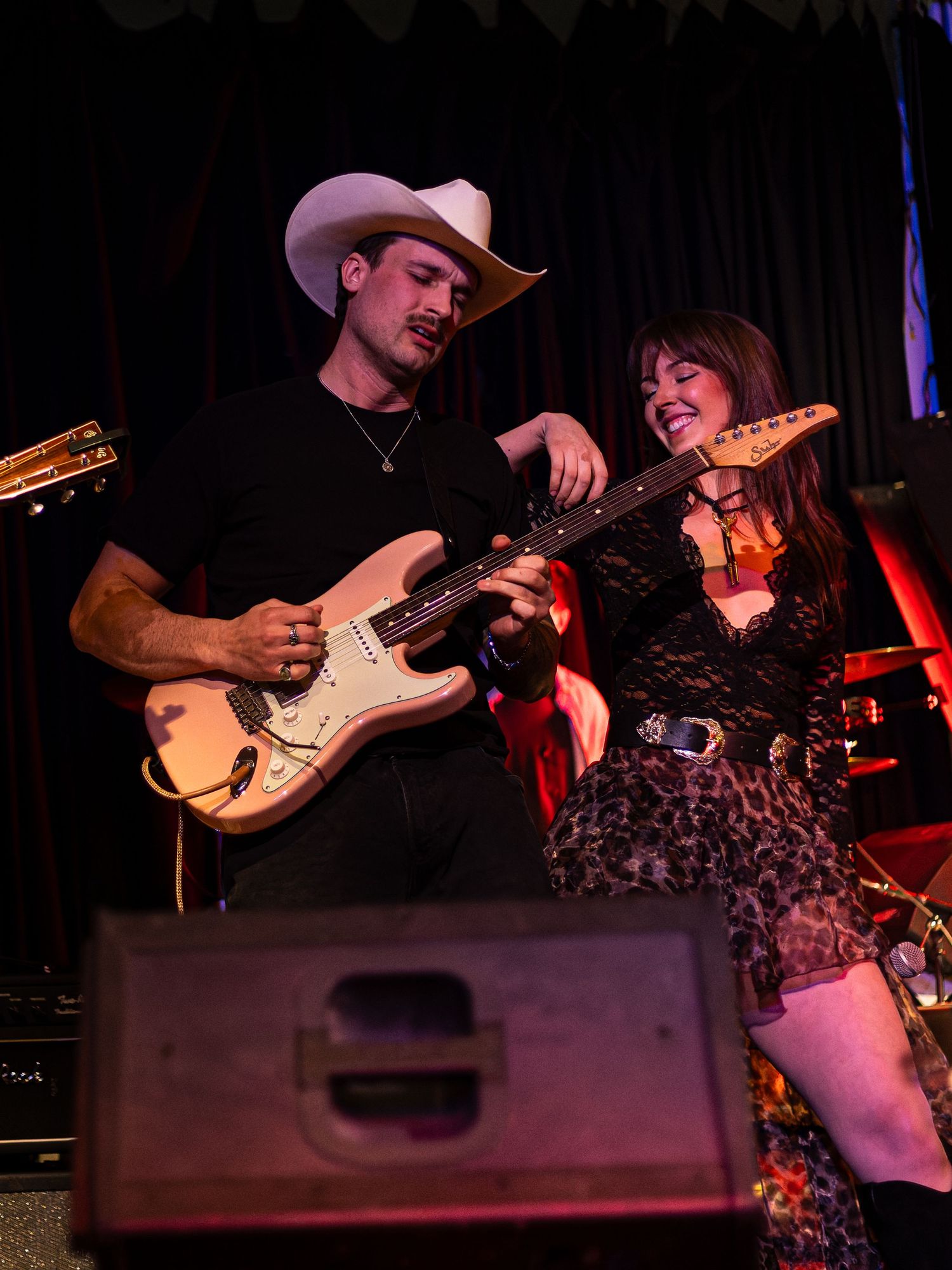 A man in a white cowboy hat plays electric guitar on stage while a woman in leopard print looks on smiling.
