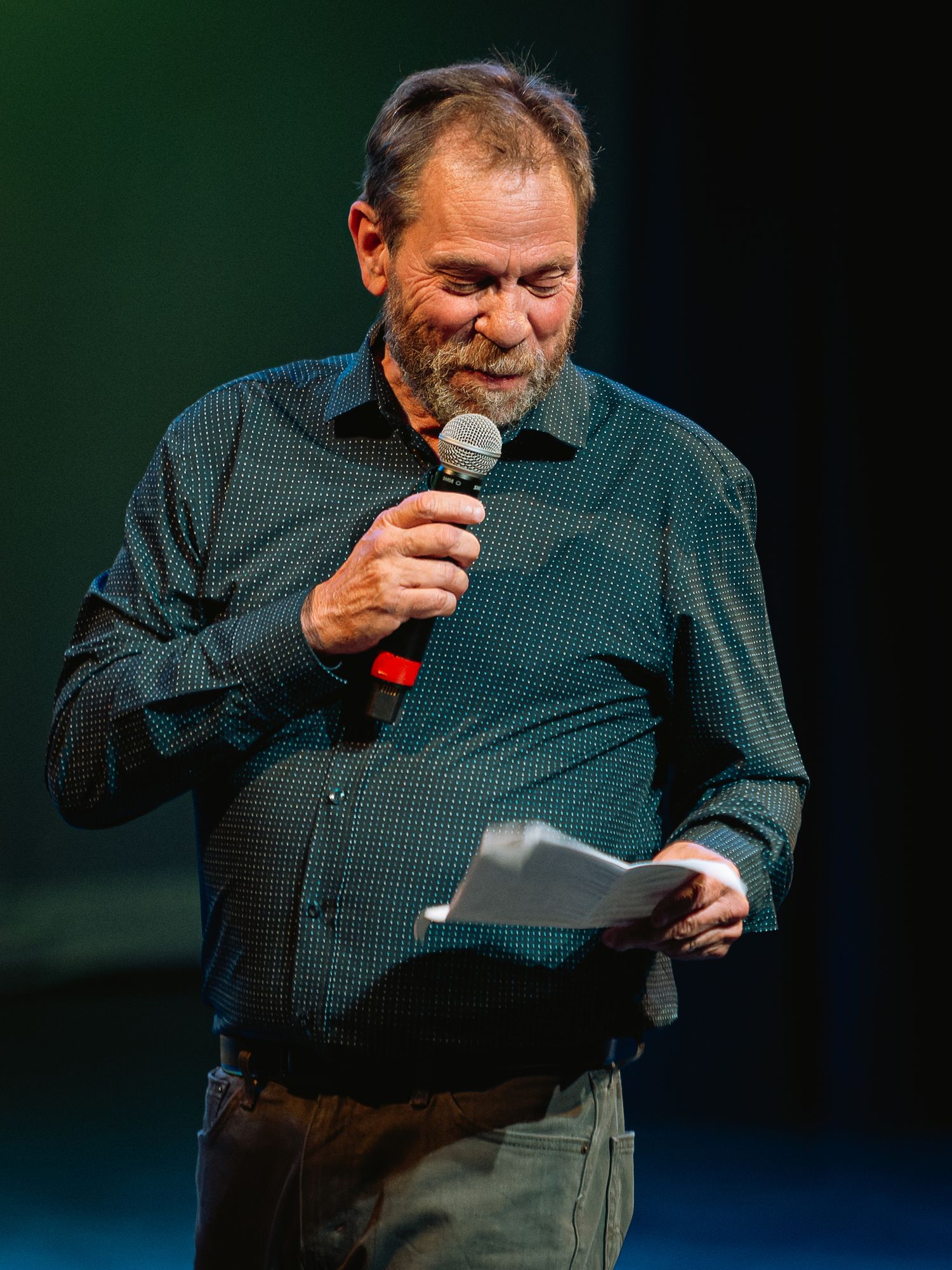 Man holding microphone and paper, speaking on stage at an event.