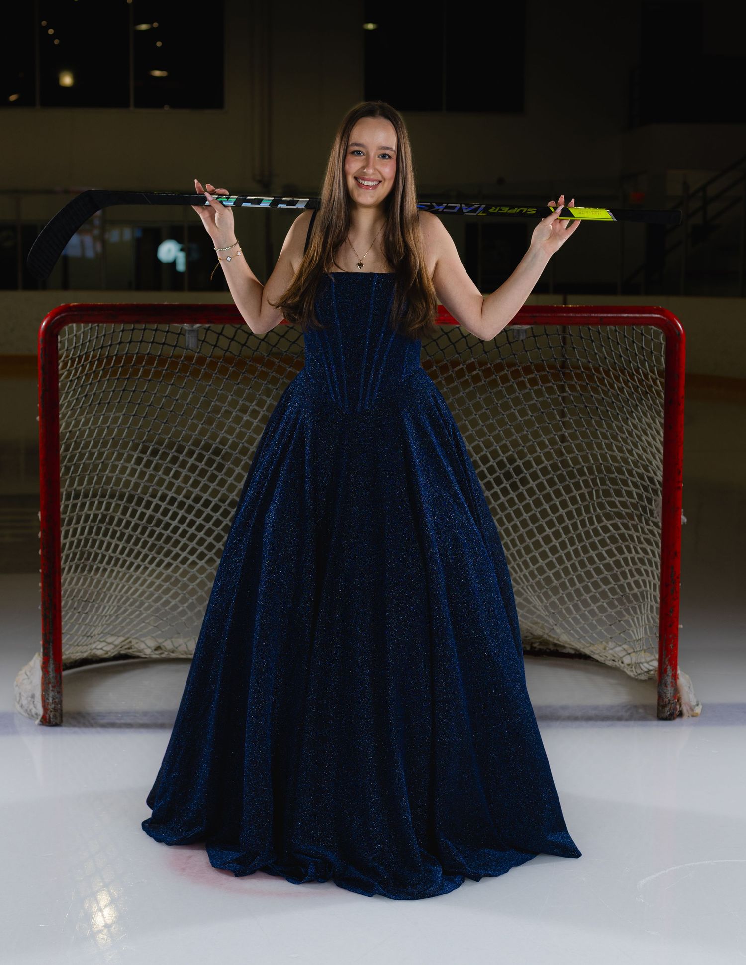 Young woman in navy blue ball gown holding hockey sticks poses in front of a hockey net on ice rink.