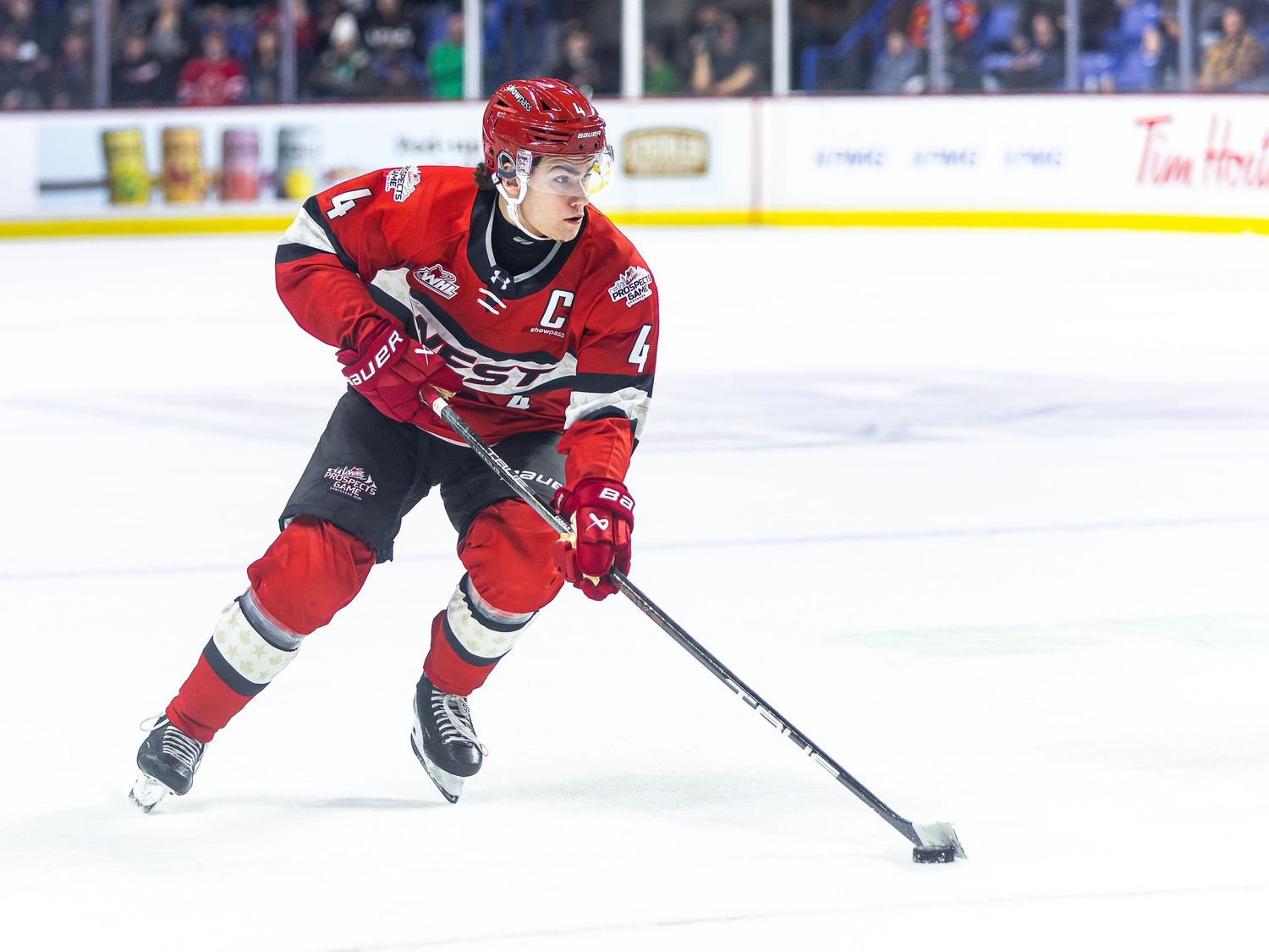 Hockey player in red uniform with captain C patch skating with puck during WHL game on indoor ice rink.