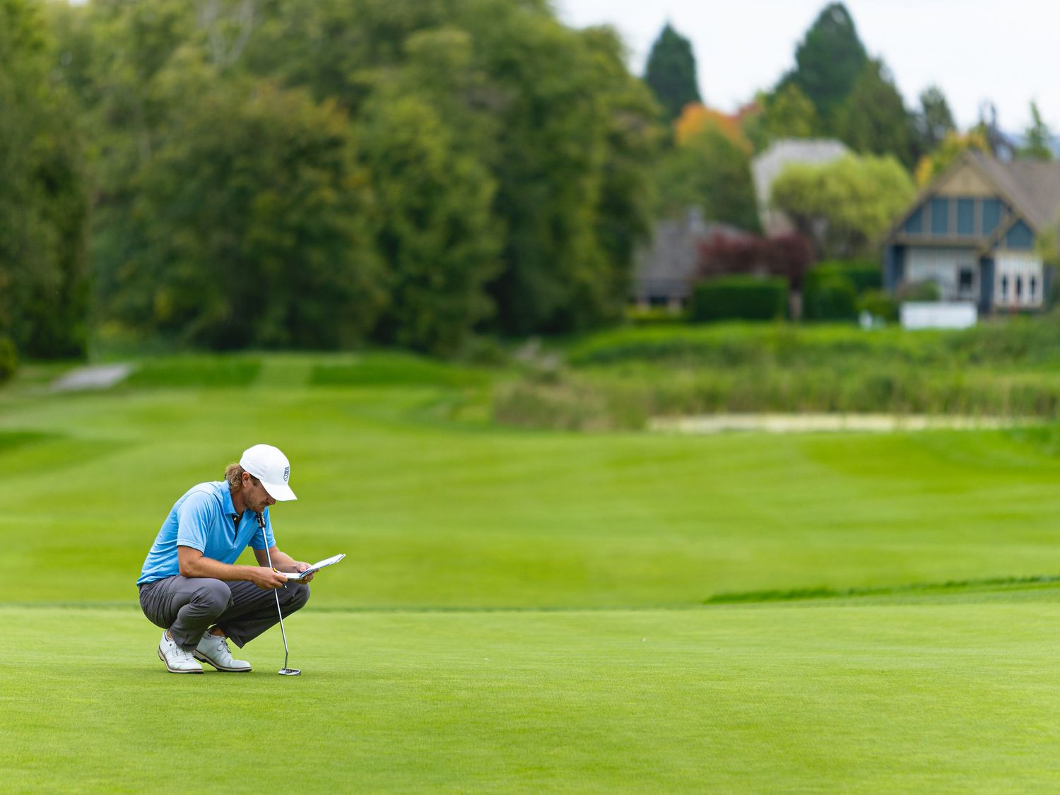 Golfer crouching on lush green fairway, analyzing putt line during a golf tournament with trees and clubhouse behind.