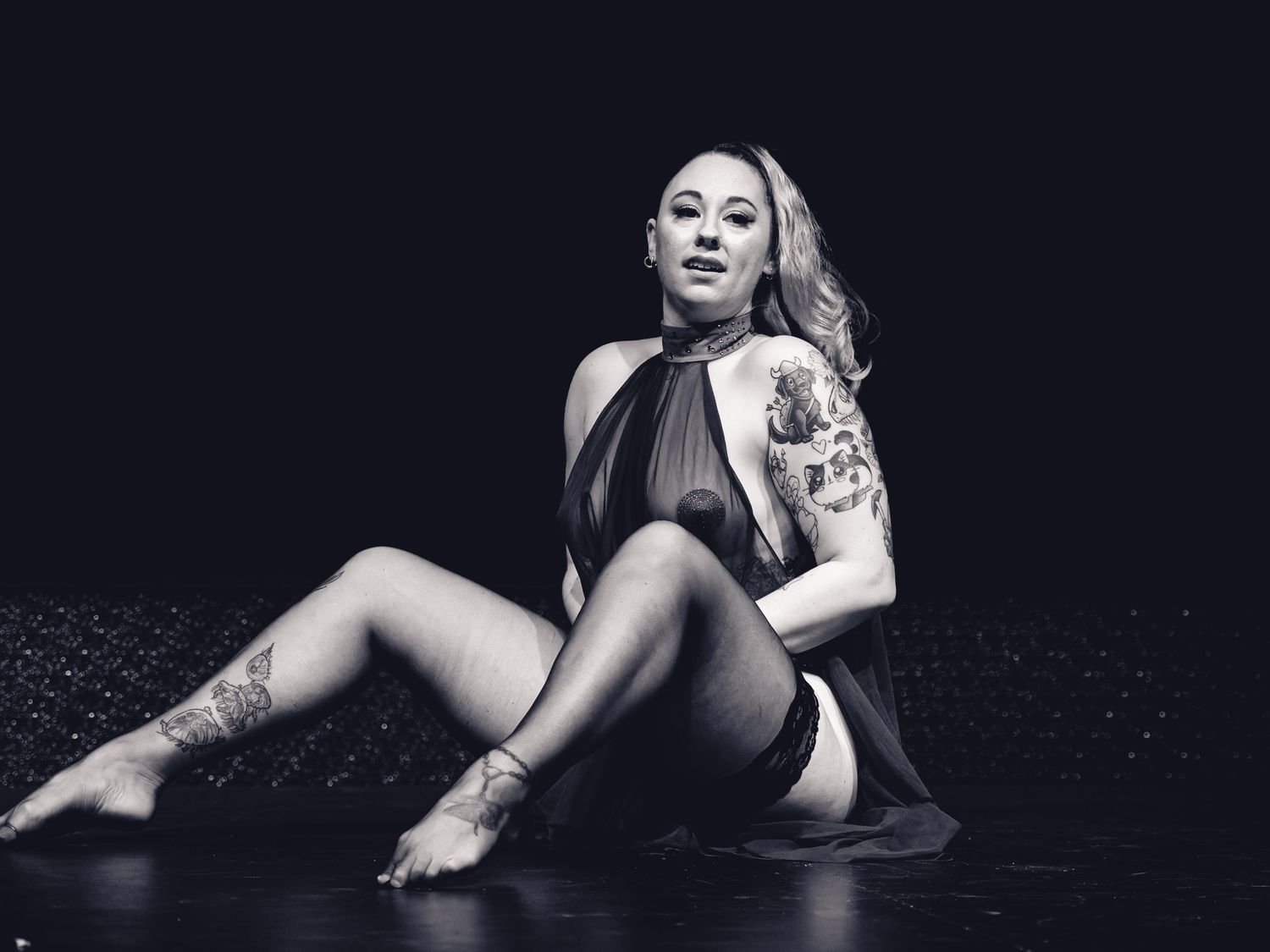 Black and white photo of a tattooed woman in a sheer bodysuit posing gracefully on a dark stage floor.