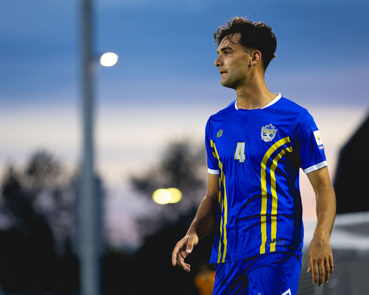 Male soccer player in blue and yellow number 4 jersey stands on field at dusk.