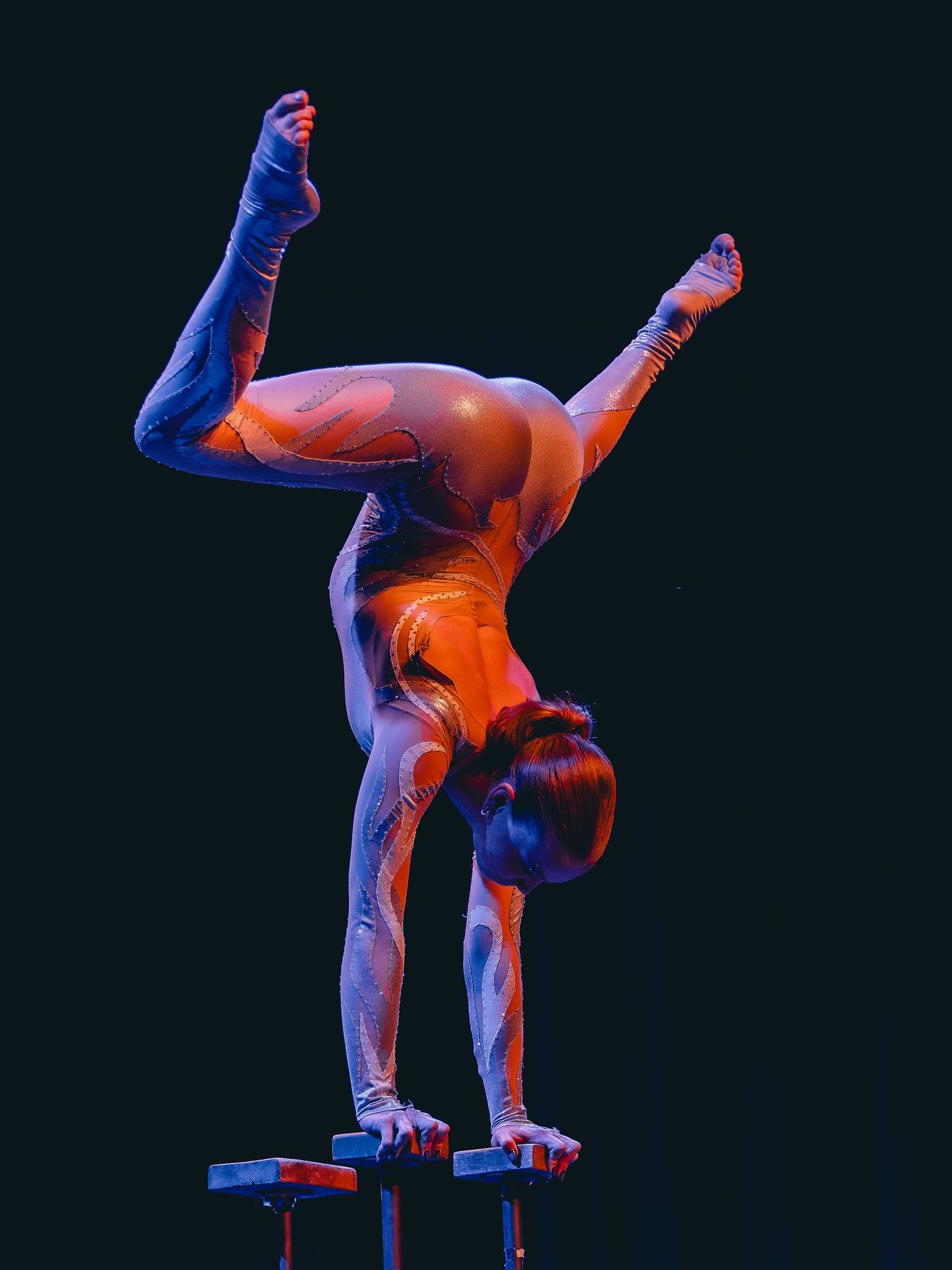 Acrobat performing a handstand on parallel bars under dramatic red and blue stage lighting against black background.