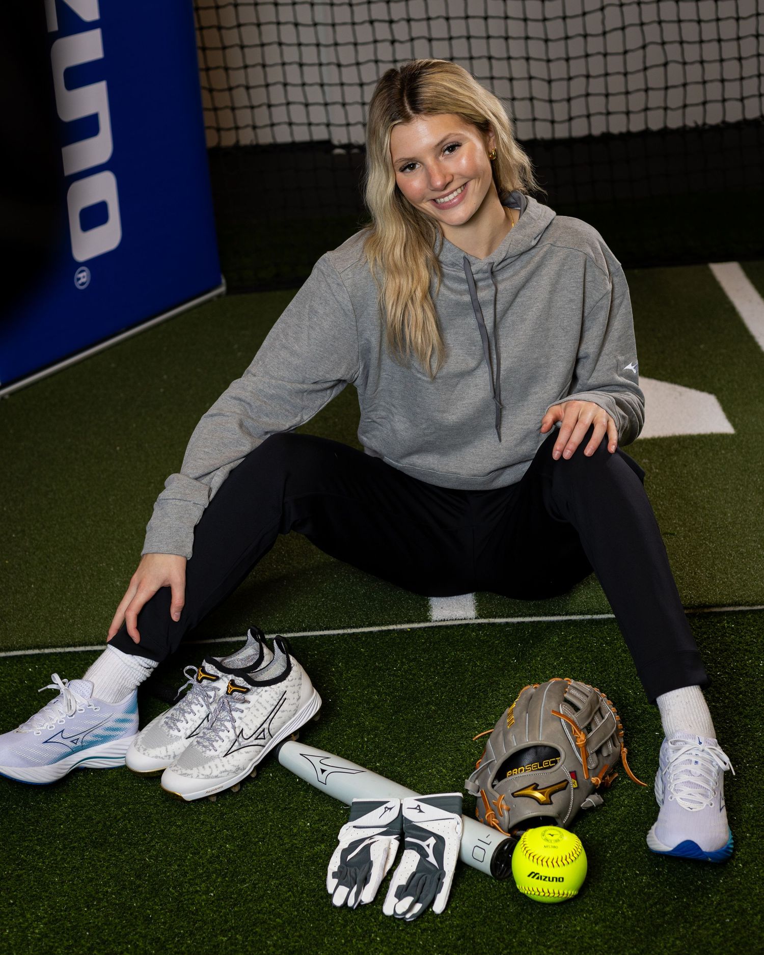 Female softball player poses with Mizuno gear, glove, cleats, and yellow softball on indoor turf.