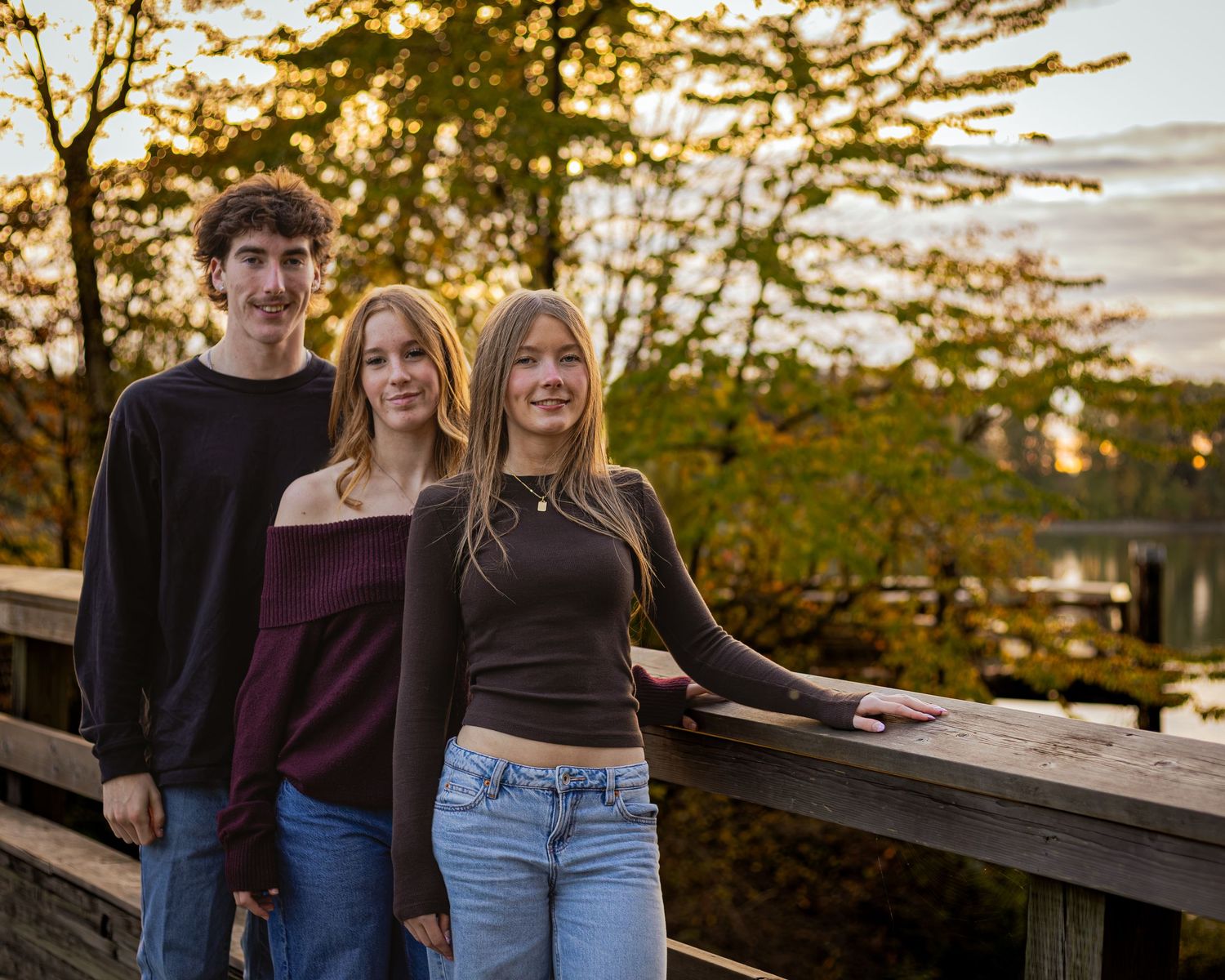 Three young adults pose on a wooden bridge surrounded by autumn trees at golden hour near a serene lake.