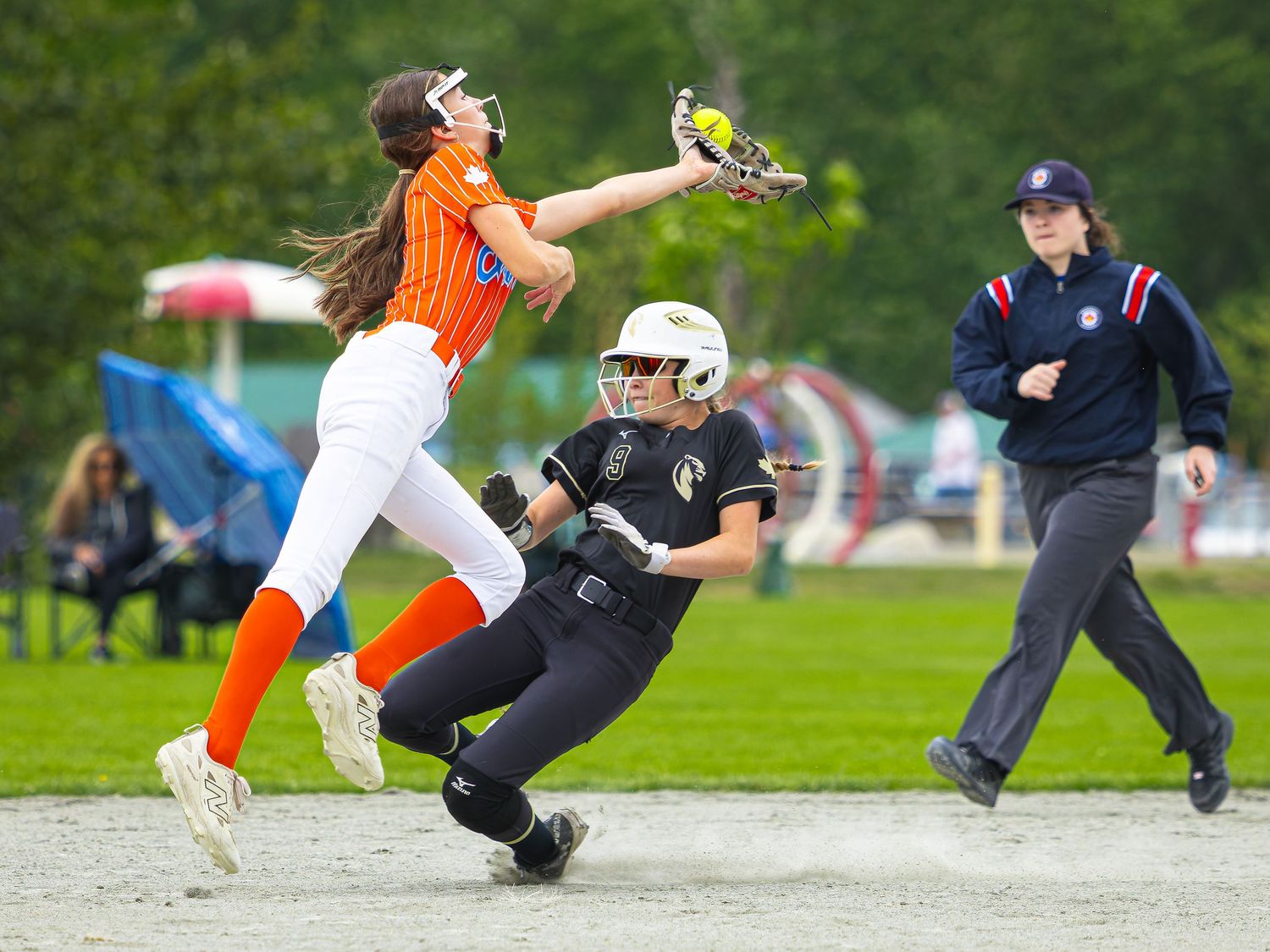 Softball player leaps to catch ball while opponent slides into base during competitive game with umpire watching.