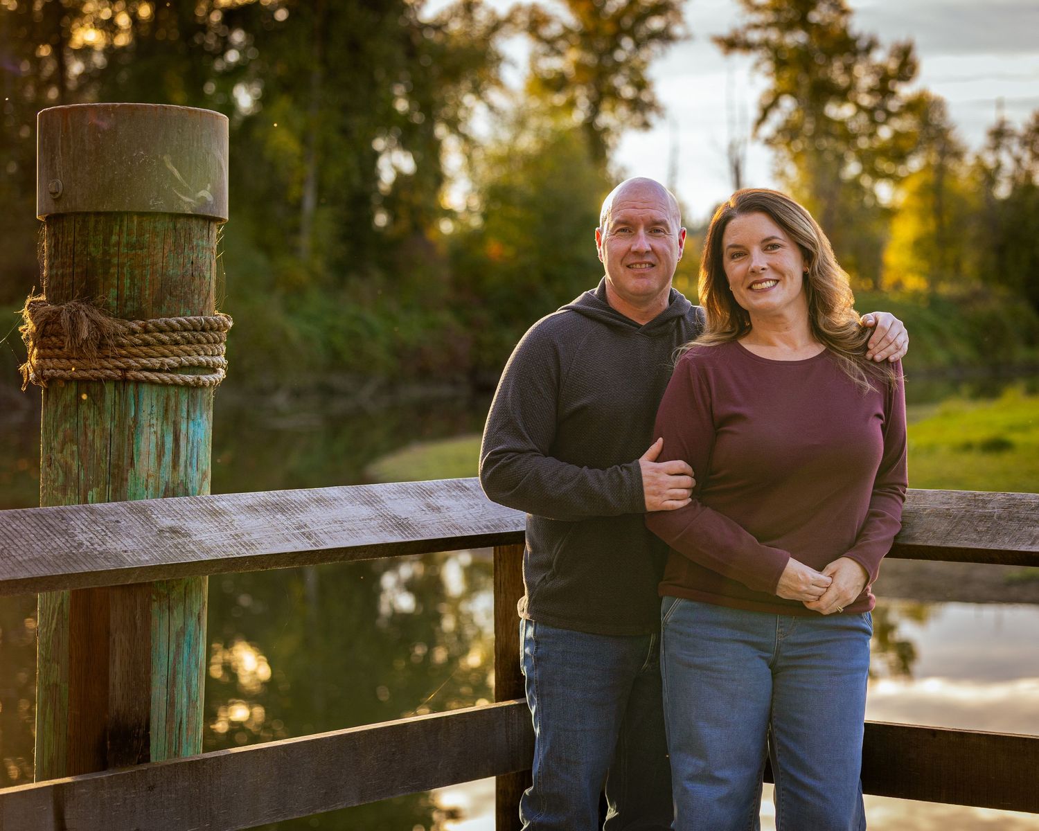 Smiling couple posing together on a wooden bridge surrounded by lush green trees and a serene river backdrop.