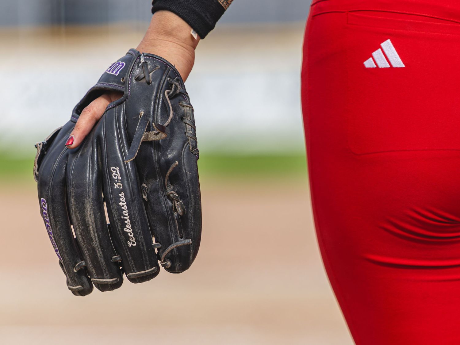 Close-up of a softball player wearing red Adidas pants holding a black Rawlings baseball glove on a dirt field.