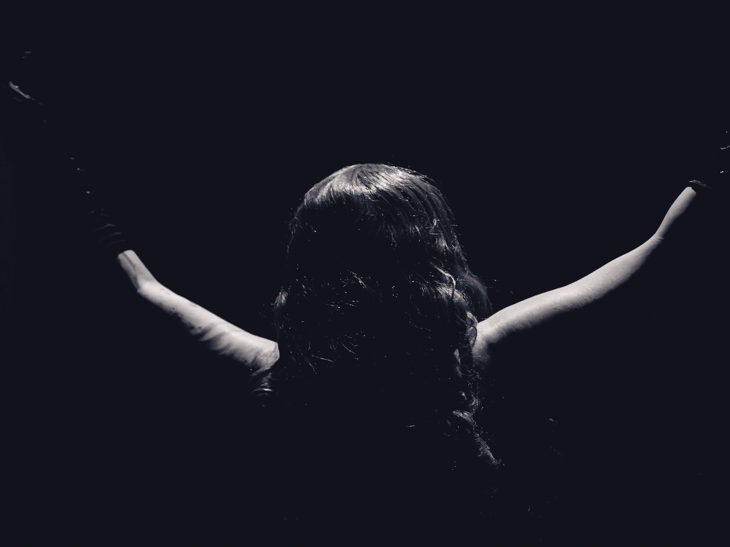 Black and white photo of a person with arms raised wide against a dark background, viewed from behind.
