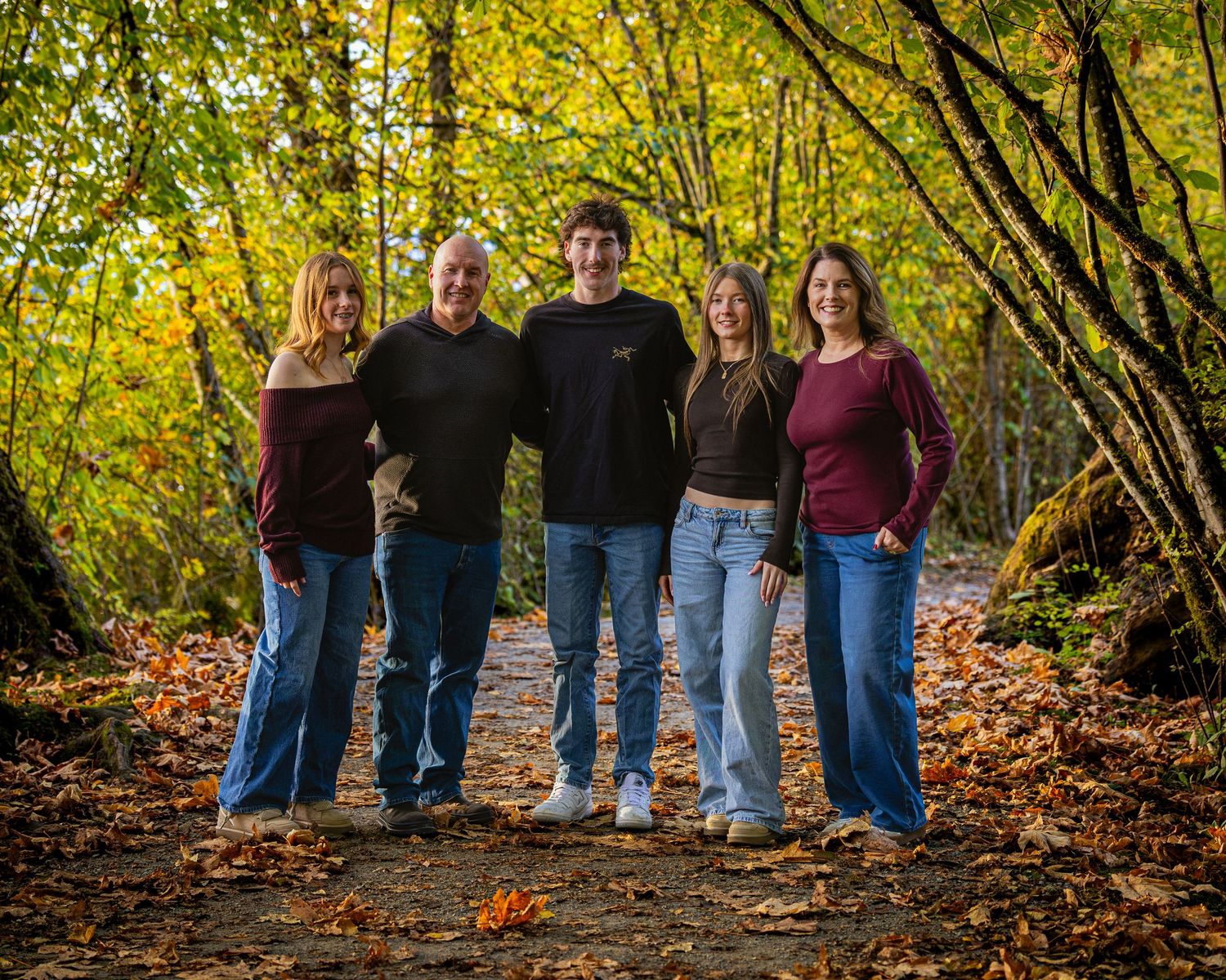 Five people pose together on a leaf-covered forest path surrounded by vibrant autumn foliage.
