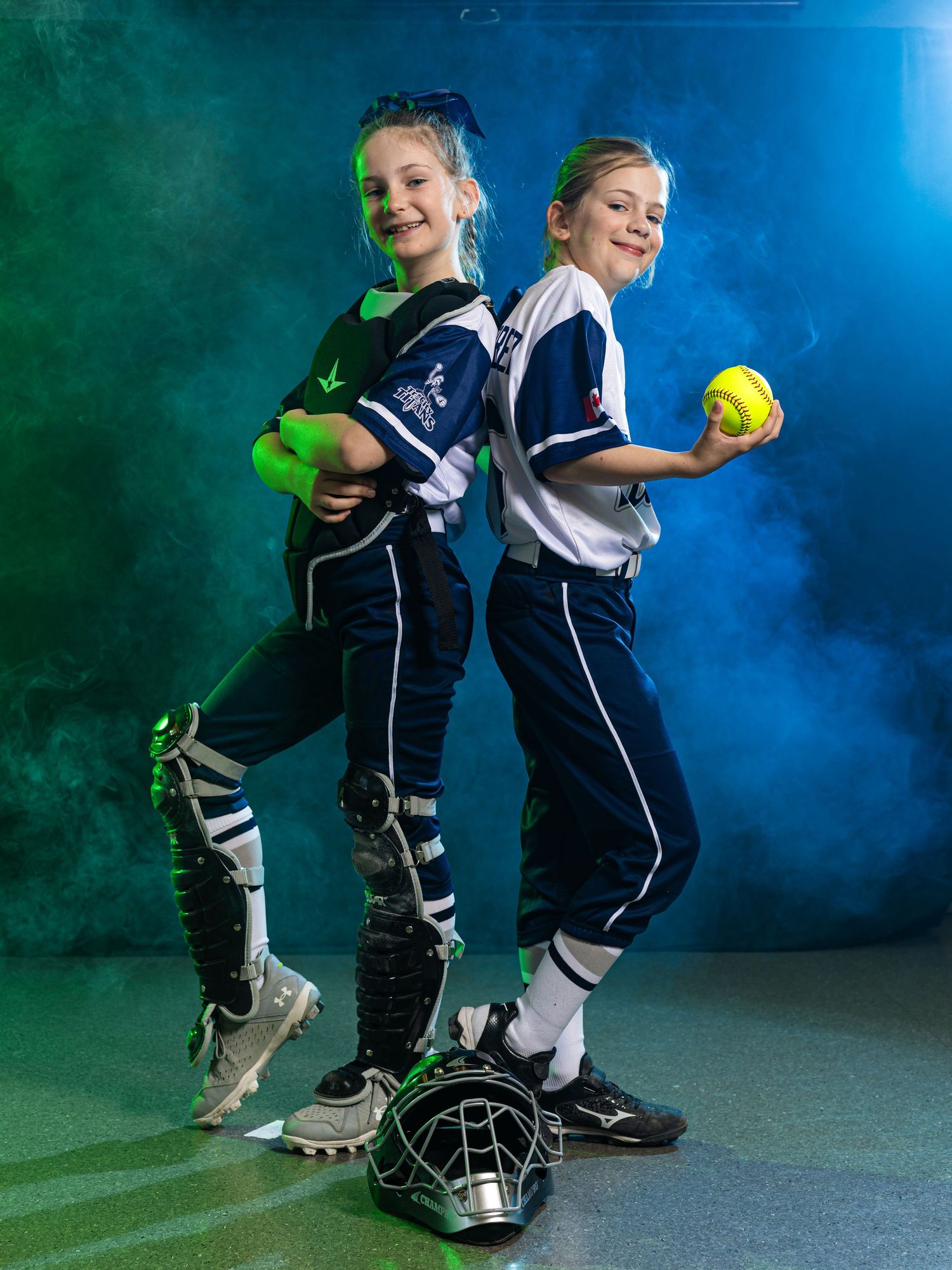 Two young girls in softball uniforms pose back-to-back, one holding a yellow ball, with dramatic teal and green smoke background.