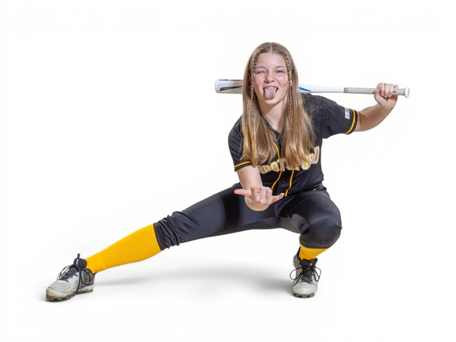 Young female softball player in black and gold uniform poses dynamically with bat behind shoulders, sticking out tongue.