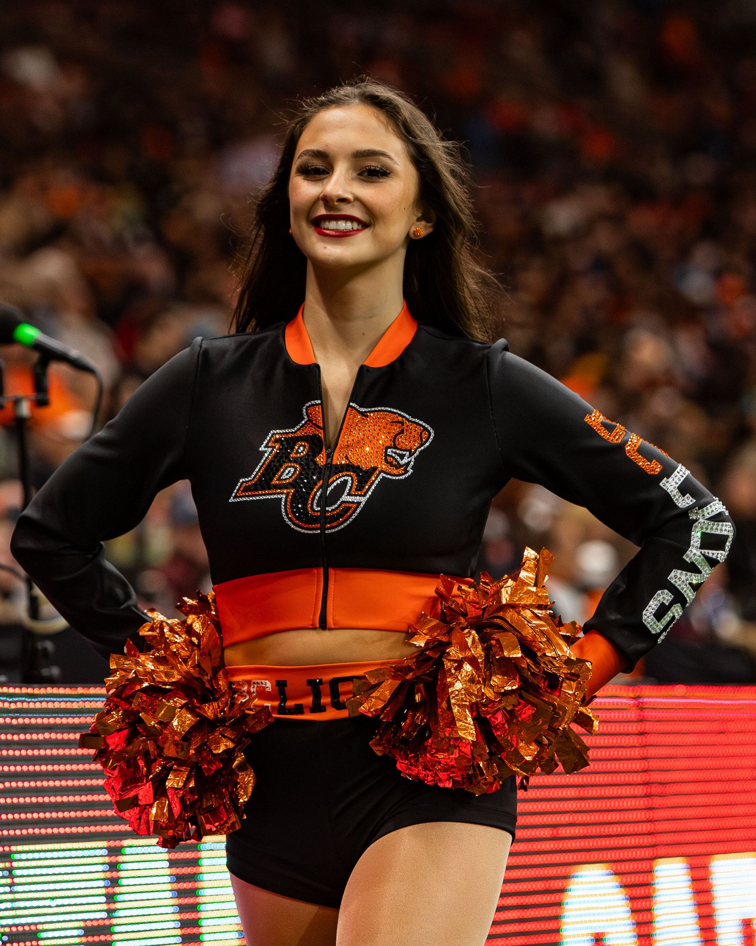 BC Lions cheerleader smiling in black and orange uniform with pom-poms at a CFL game.
