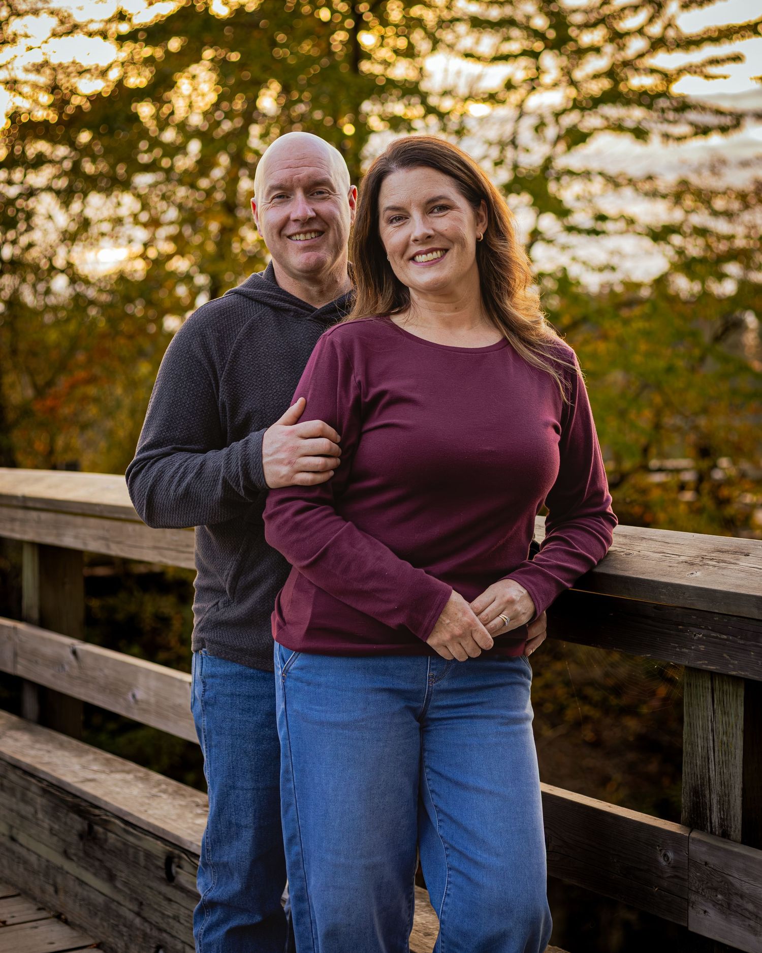 A smiling couple poses on a wooden bridge surrounded by autumn foliage in a warm outdoor portrait.