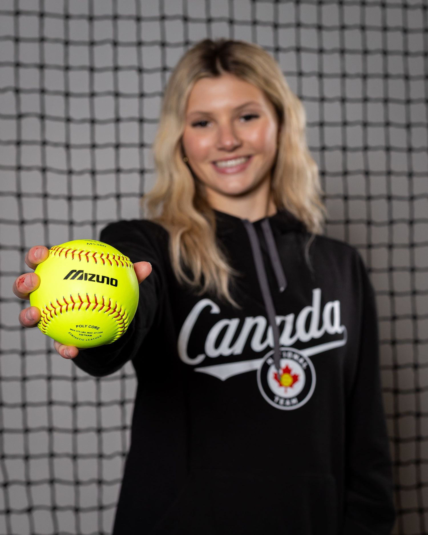 Young blonde woman in a Canada softball team hoodie holds a yellow Mizuno softball toward the camera, smiling.