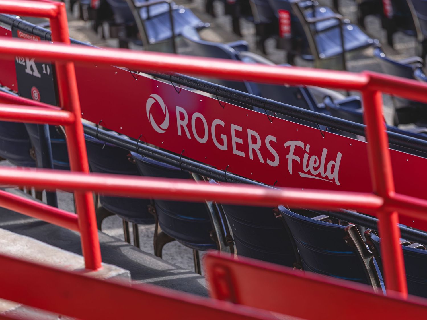 Red seats and railings at Rogers Field stadium with branded signage visible in the foreground.