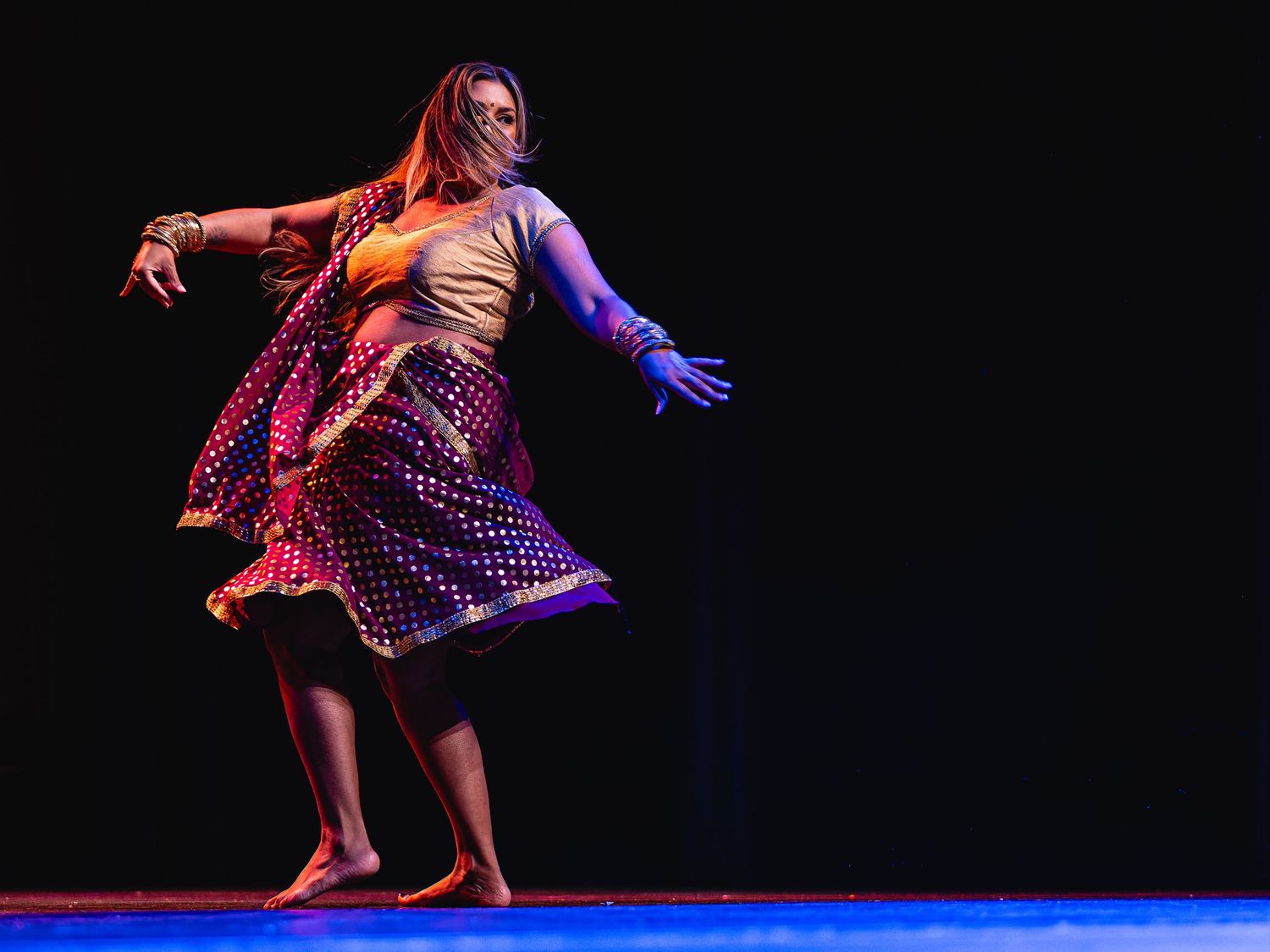 A woman performs Indian classical dance on stage, wearing a colorful polka-dot skirt and traditional jewelry.