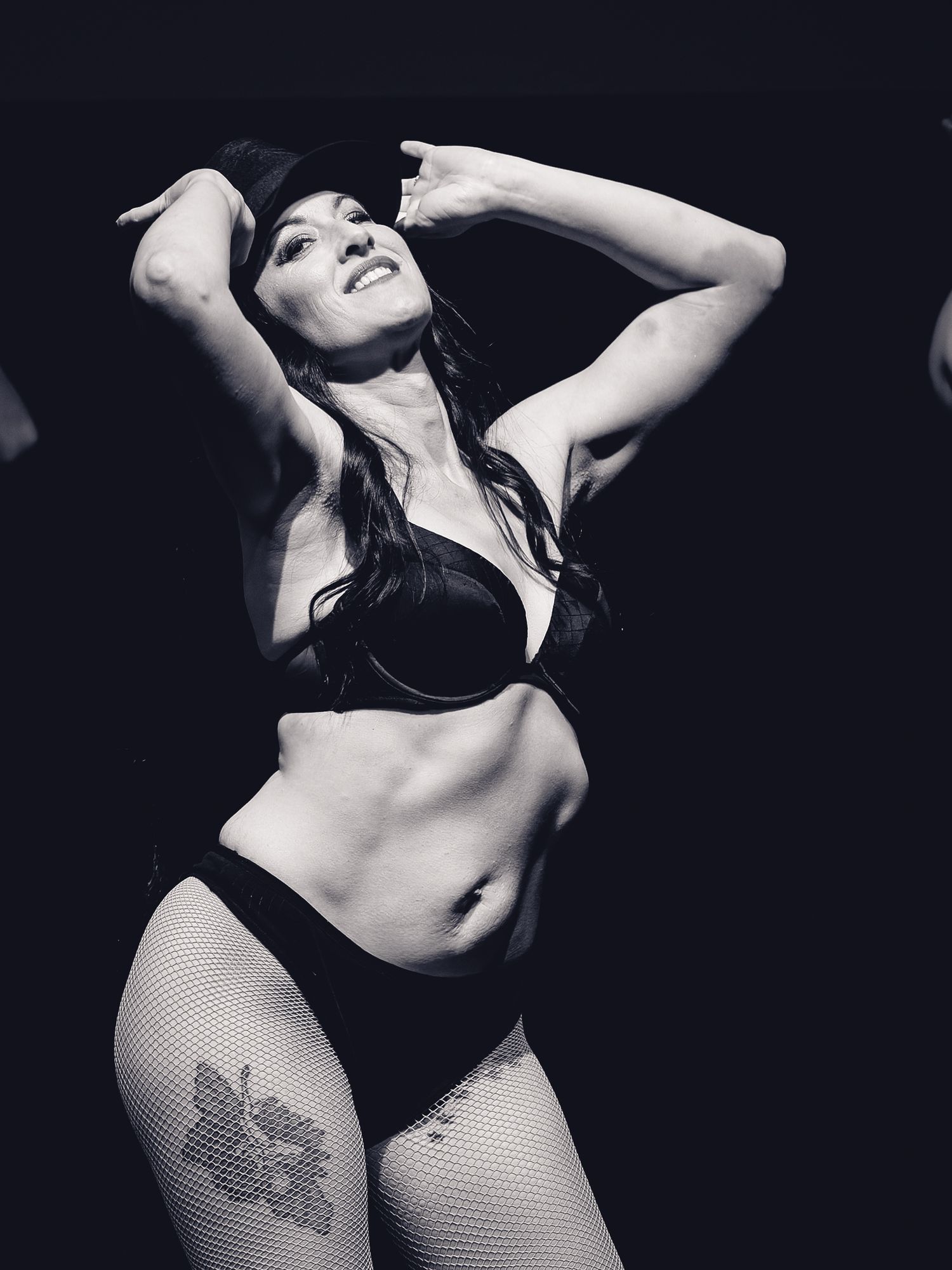 Black and white photo of a woman posing confidently in lingerie and fishnet stockings under dramatic stage lighting.