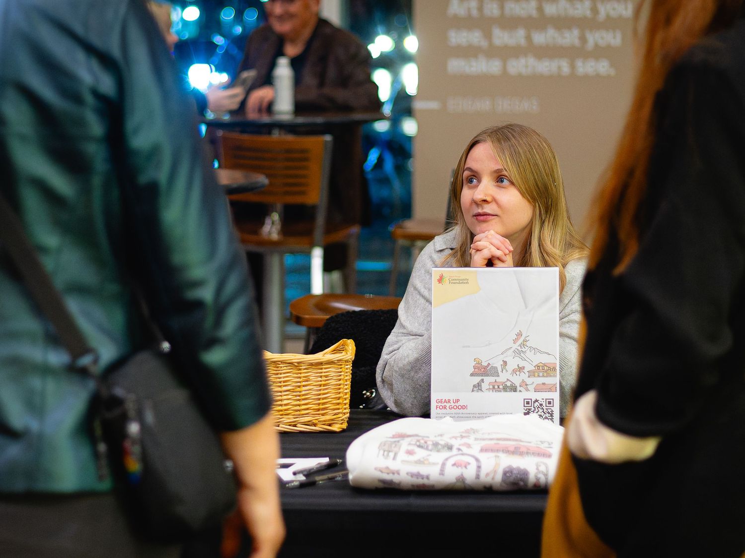 Woman sitting at an event table with a basket and printed materials, engaged in conversation at an indoor venue.