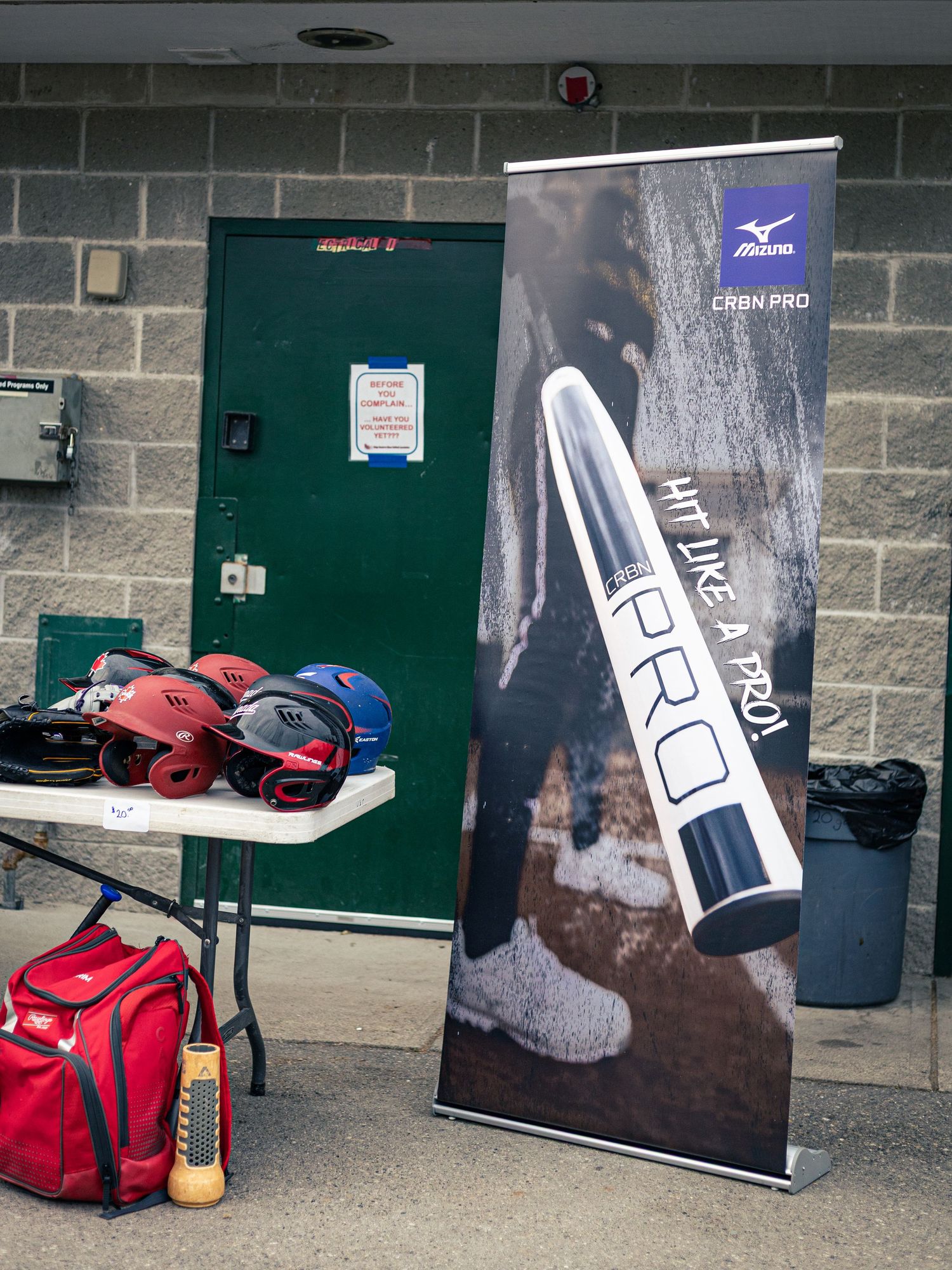 Mizuno CRBN PRO baseball bat banner display beside helmets and equipment bag at a baseball facility.