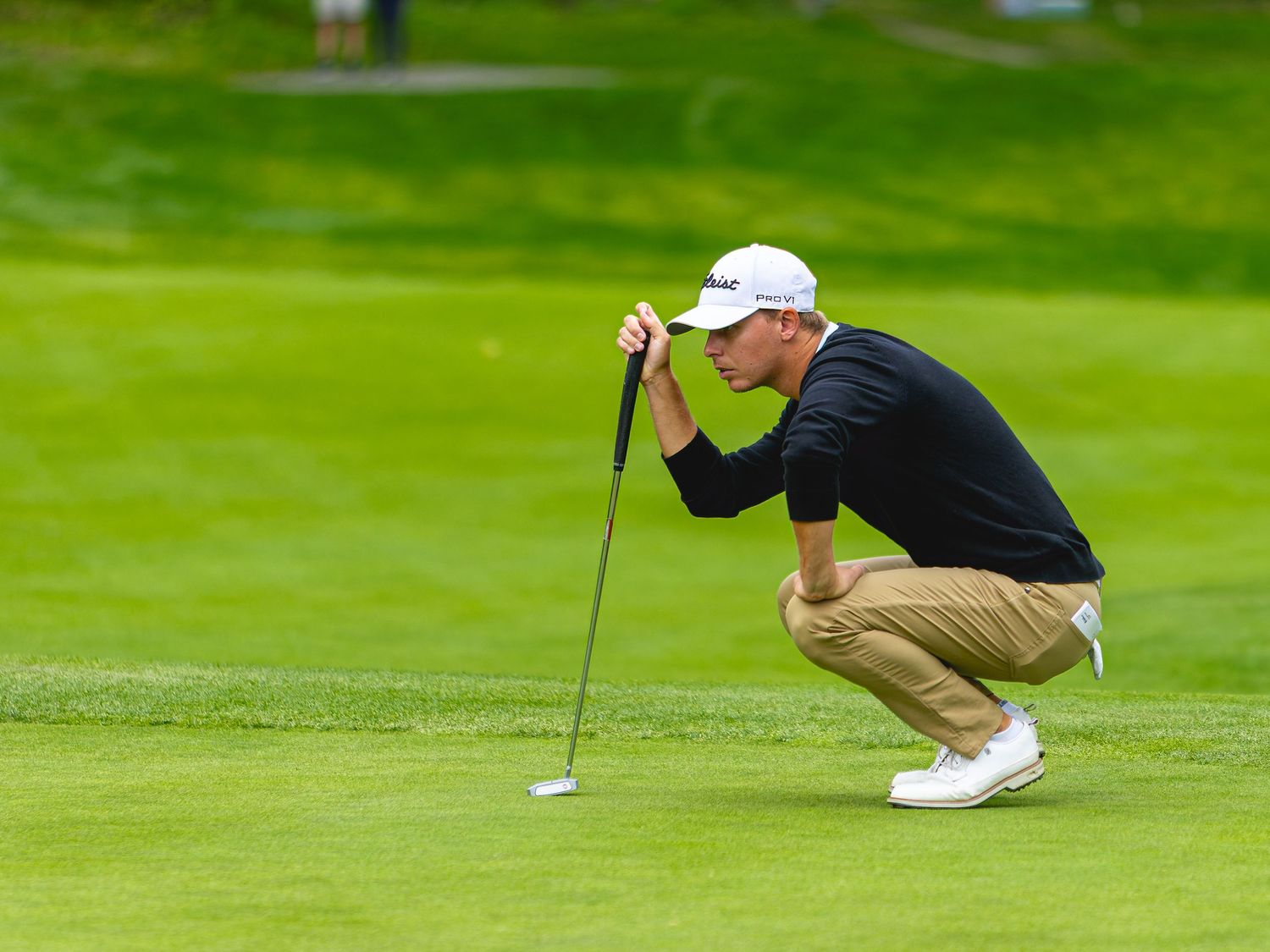 Golfer crouching on green, reading putt line while holding putter on lush golf course.