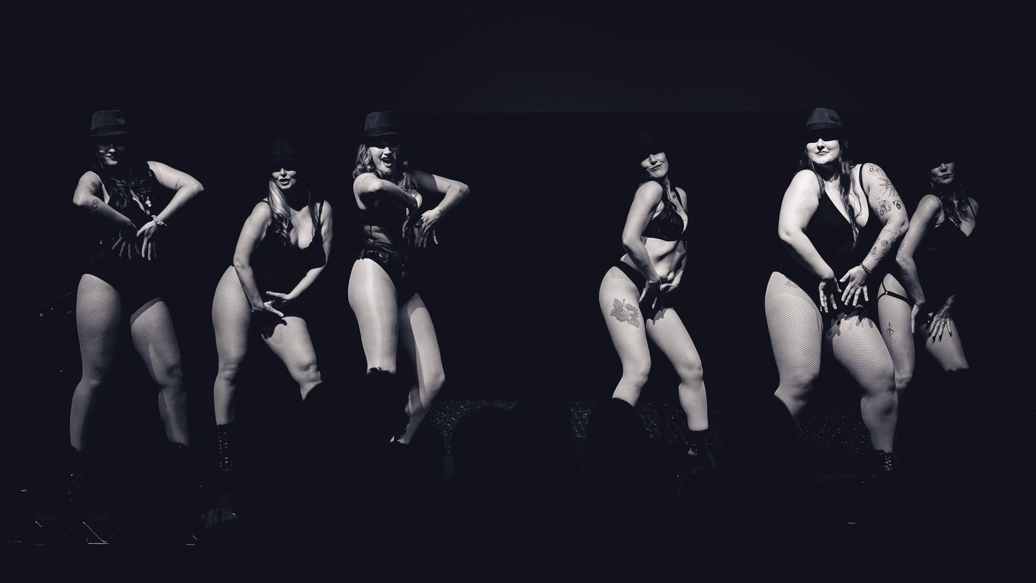 Black and white photo of five performers in black bodysuits and masks dancing on a dimly lit stage.