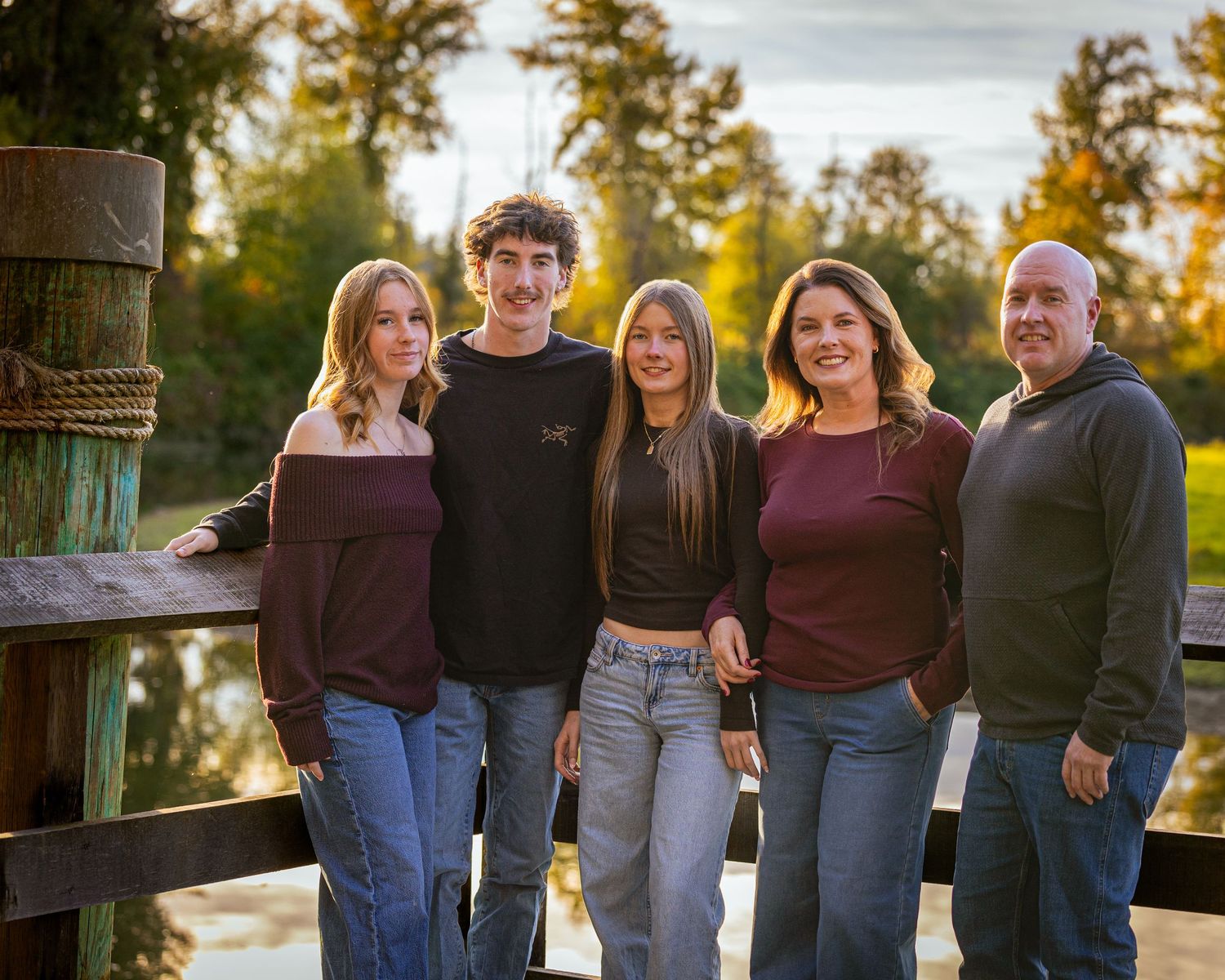 Family of five poses on a wooden dock surrounded by autumn trees in coordinating burgundy outfits.