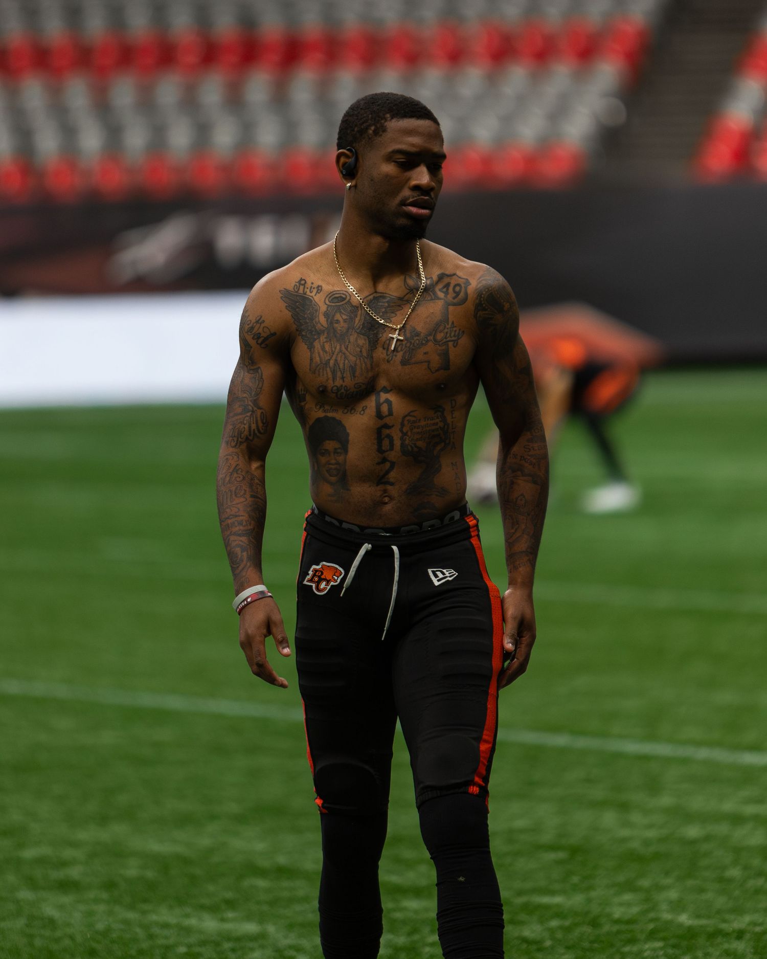 Shirtless tattooed athlete in black New Era sweatpants stands on a football field inside a stadium.