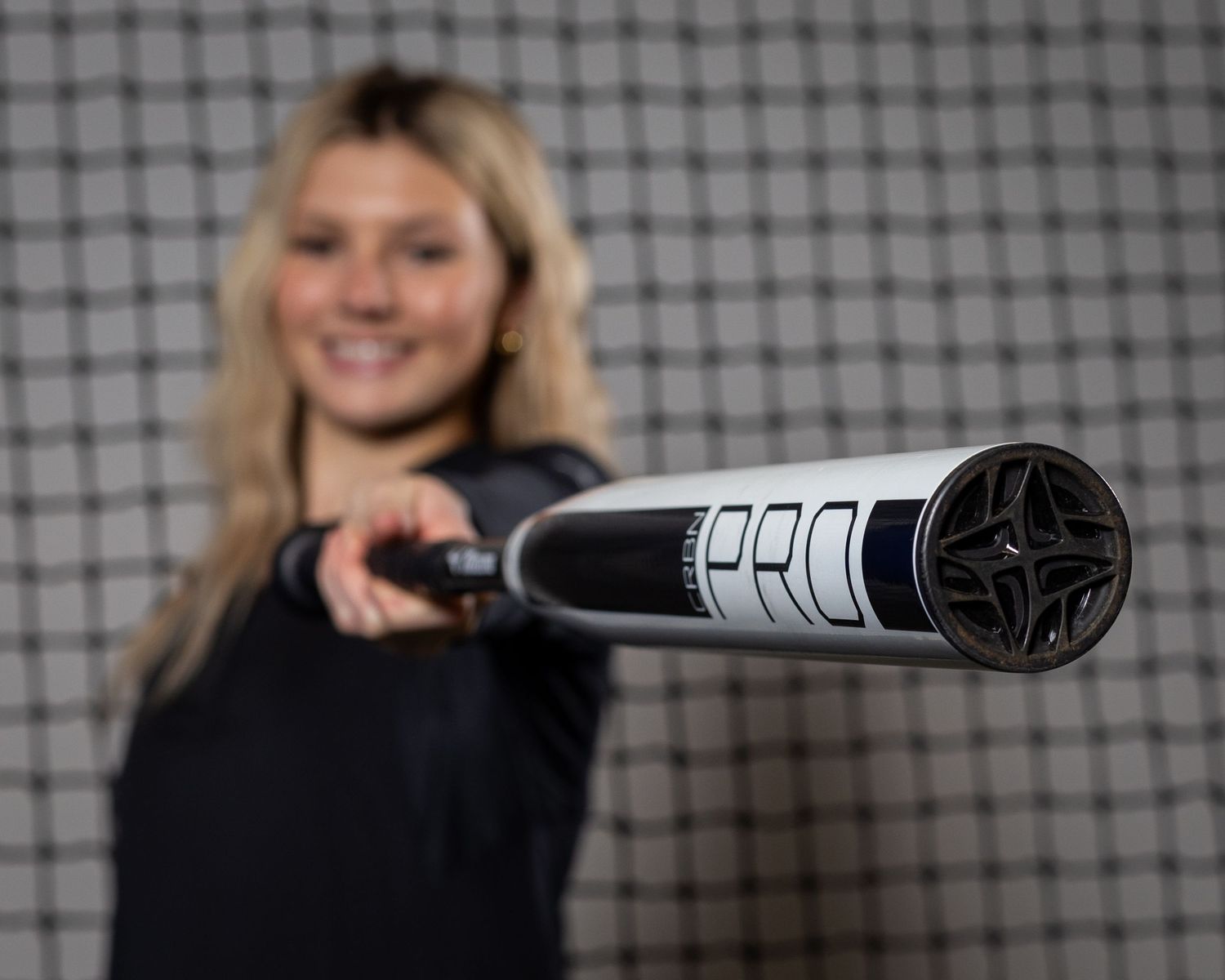 Blonde female softball player pointing a black and white PRO baseball bat toward the camera at an indoor facility.