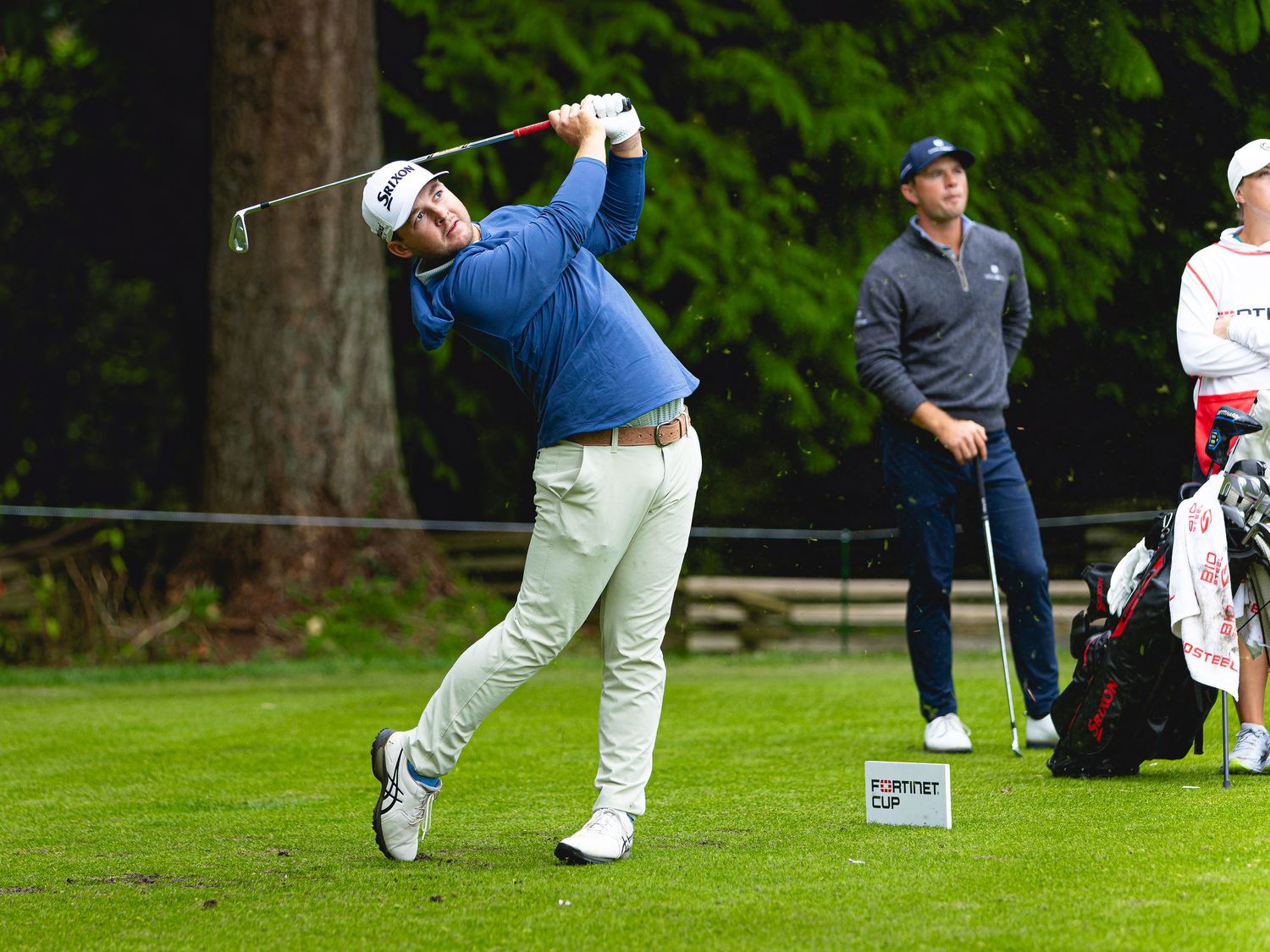 Golfer in blue shirt swings club on lush green course while spectators and caddie watch in background.