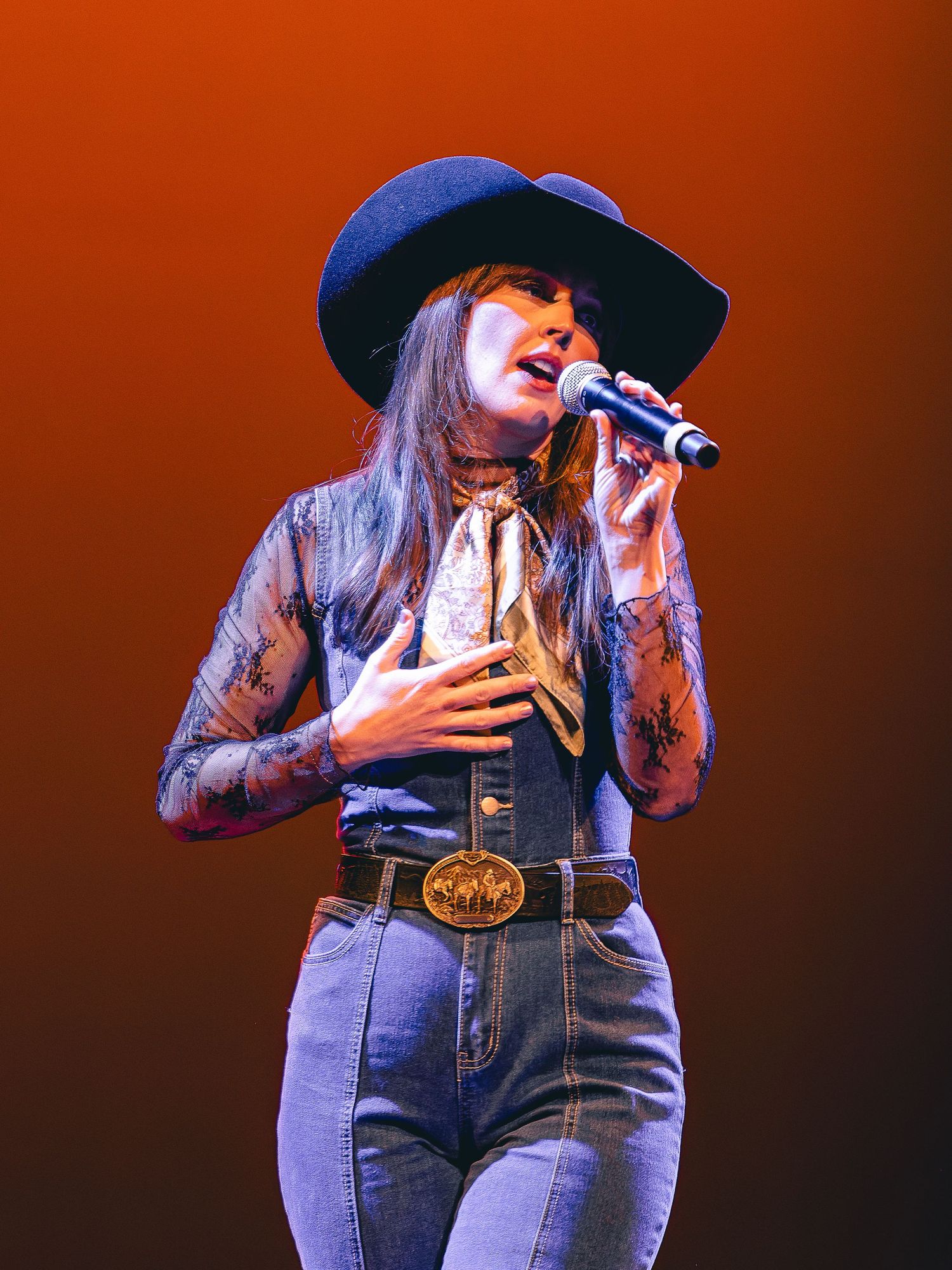 Woman singer in blue cowboy hat, floral top, and jeans with large belt buckle performs on stage with microphone.