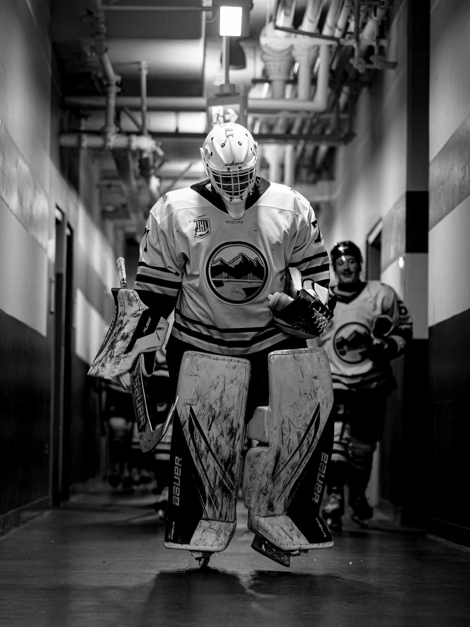 Black and white photo of a hockey goalie in full gear walking through an arena tunnel before a game.