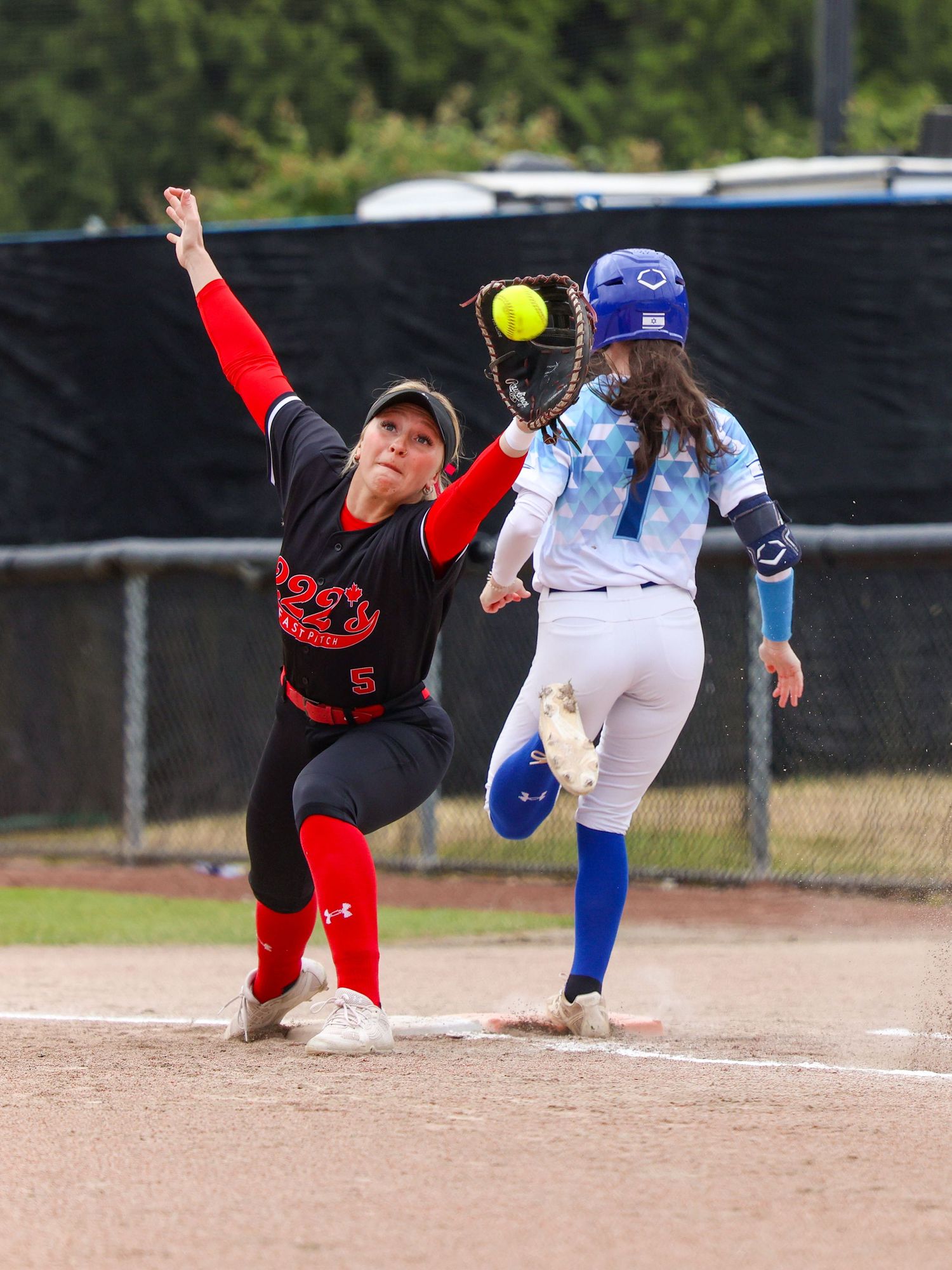 Softball player in red stretches to catch ball at first base as blue uniform runner approaches during game.