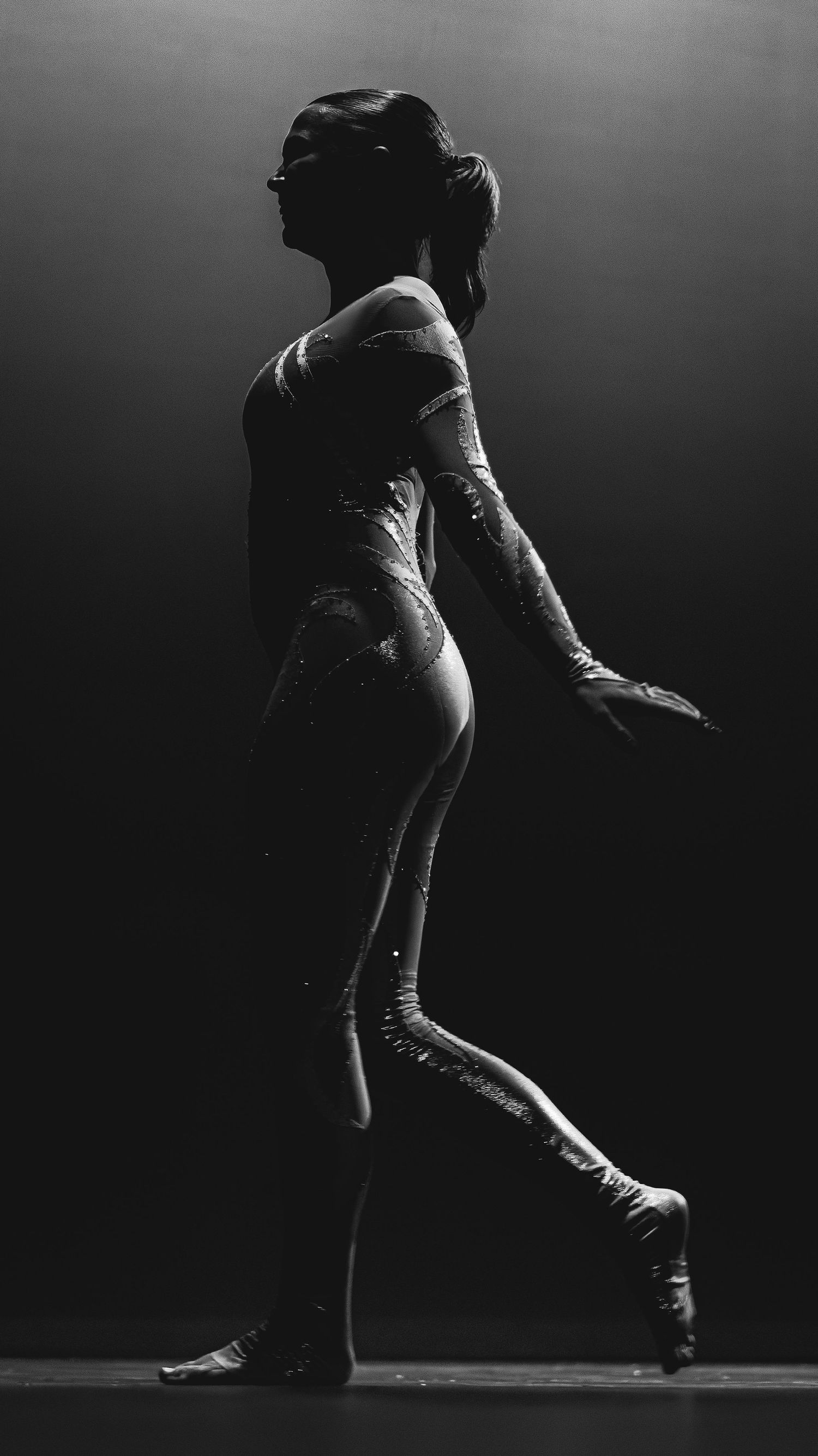 Black and white silhouette of a female dancer in a sequined leotard performing gracefully on a dark stage.