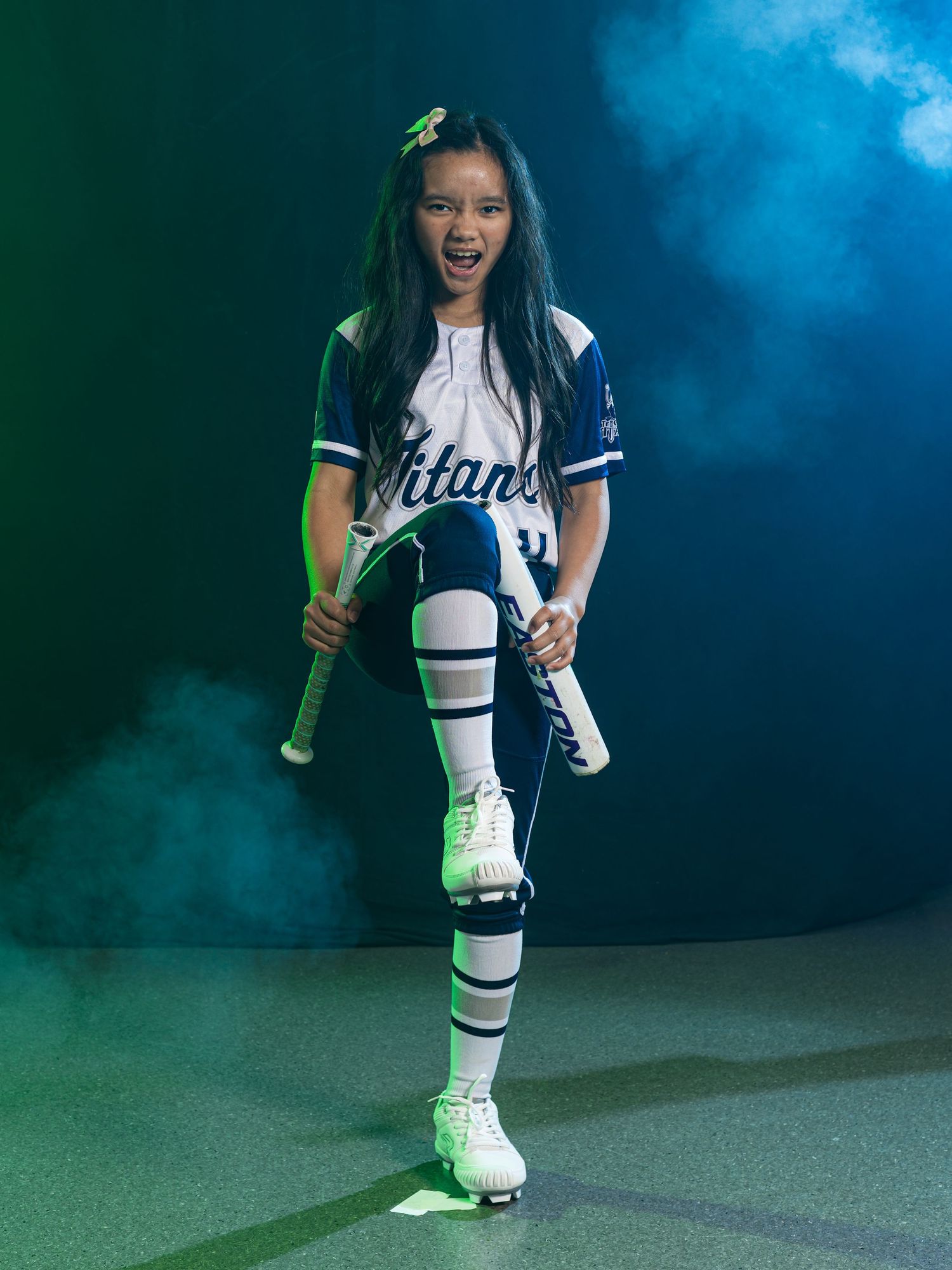 Young softball player in blue and white uniform poses confidently with bat, wearing knee-high socks and cleats.