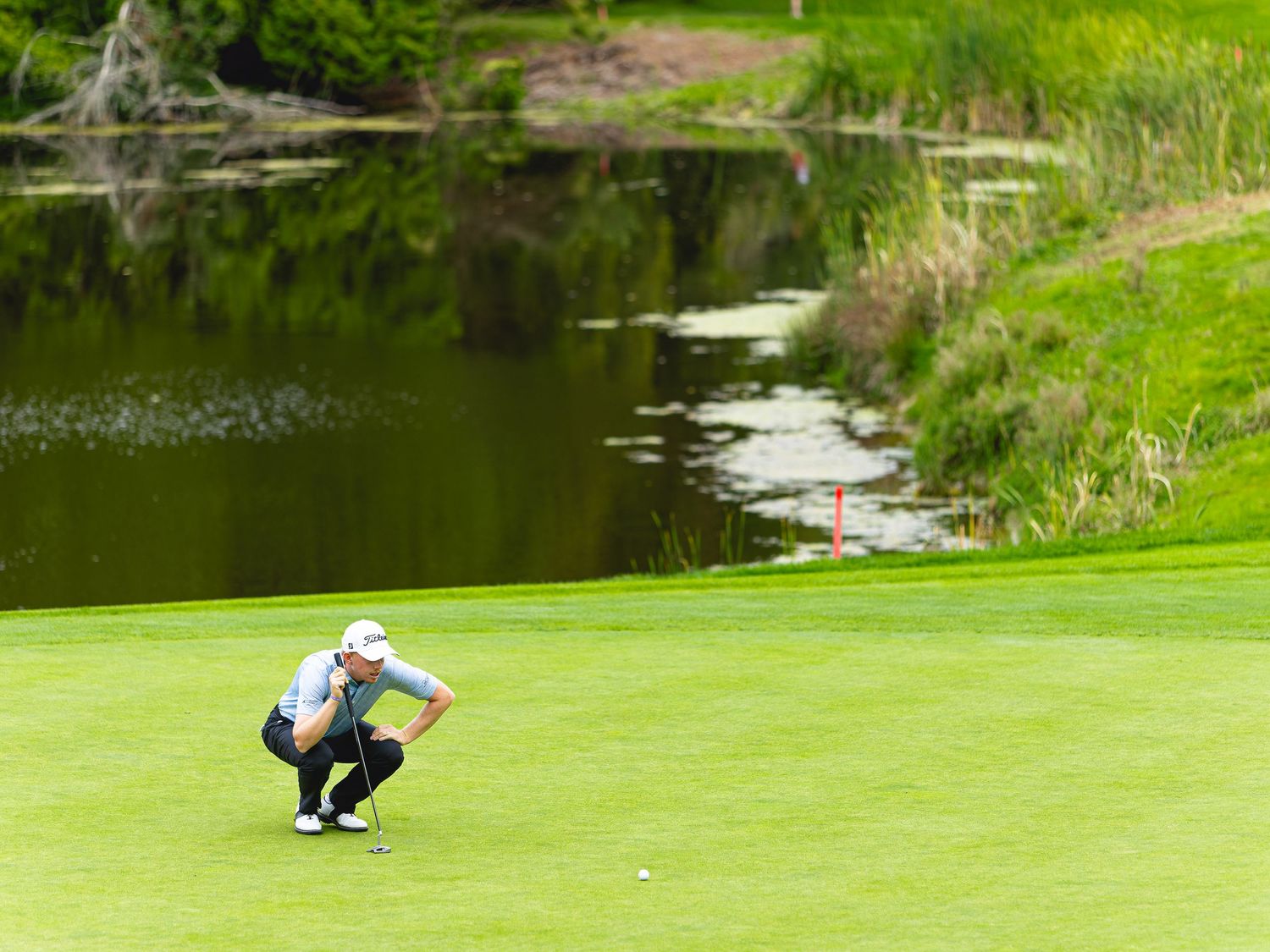 Golfer crouching on lush green putting surface near a scenic pond, reading the green with a red flag visible.