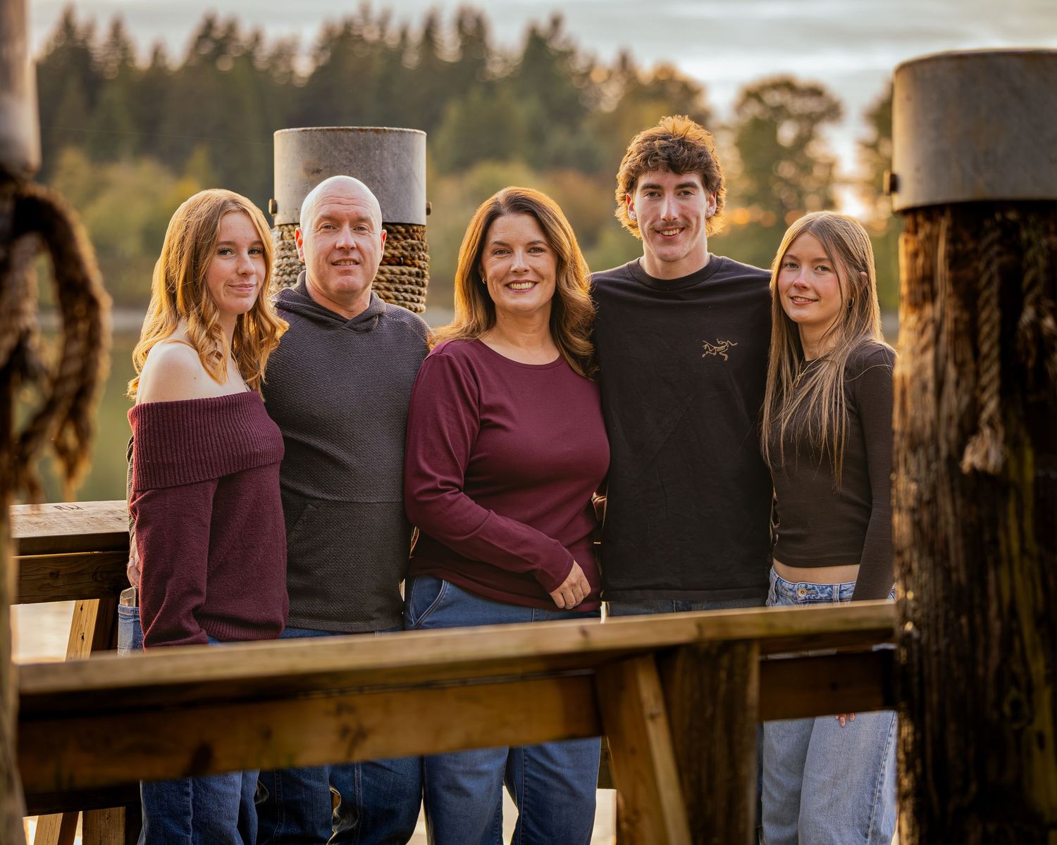 A family of five poses together on a wooden dock surrounded by trees at golden hour.