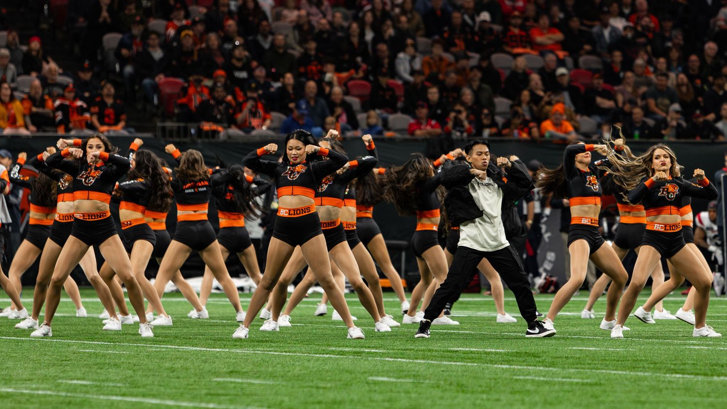 Cheerleaders in black and orange uniforms perform a synchronized dance routine on a football field during a game.