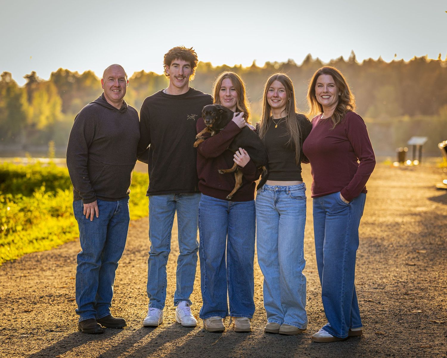 Family of five poses with a small dog on a sunlit path surrounded by golden fields at sunset.