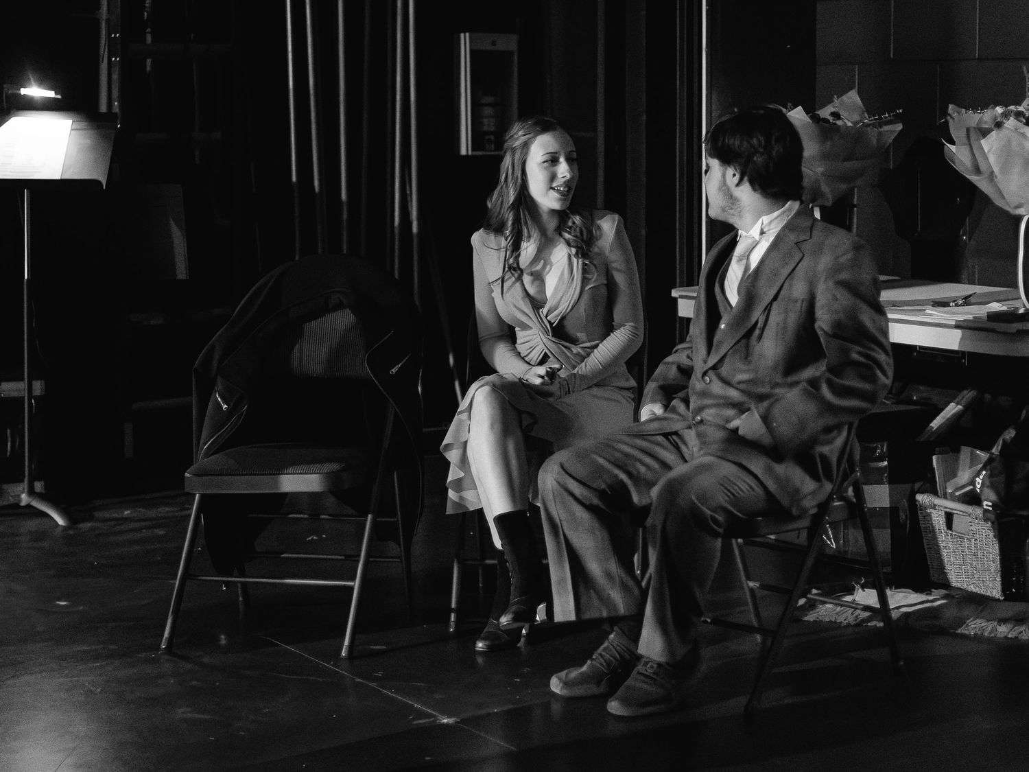 Black and white photo of a man and woman sitting on chairs conversing on a dimly lit theater stage.