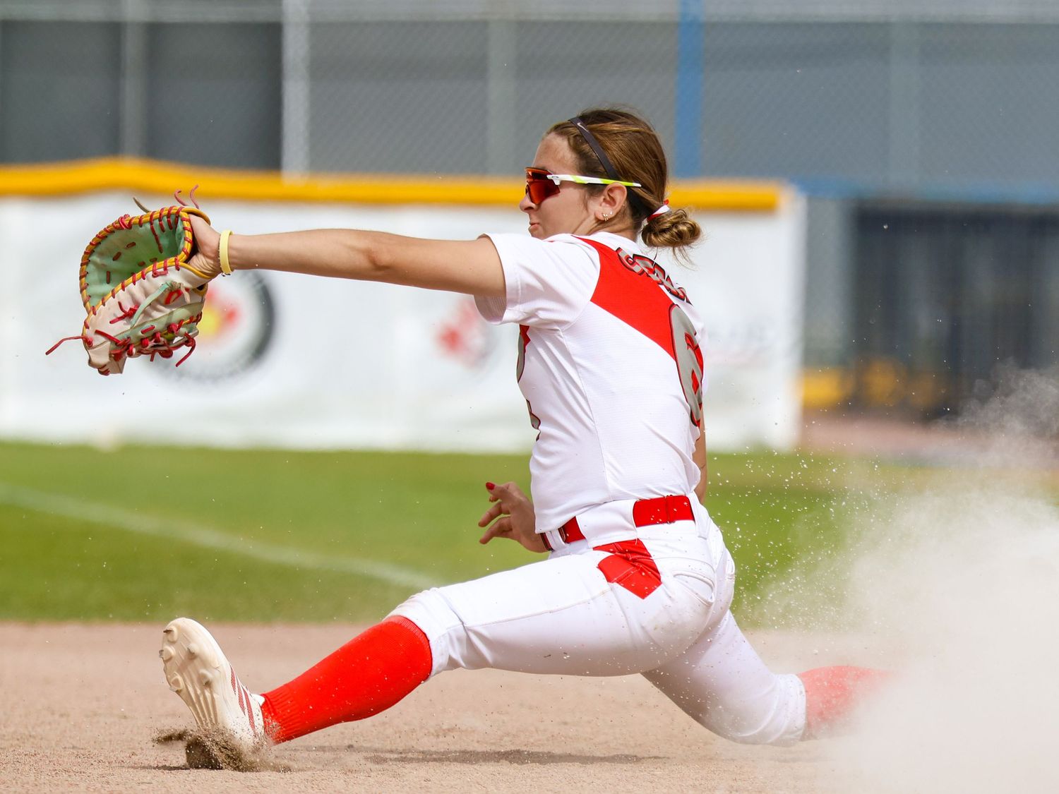 Female softball player in red and white uniform makes a dramatic fielding play, sliding in a cloud of dust near base.