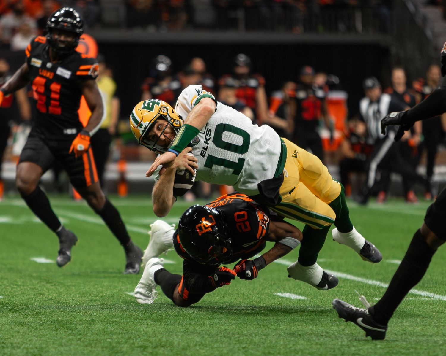 CFL football player wearing number 10 green jersey being tackled by opponent in black and orange uniform during night game.