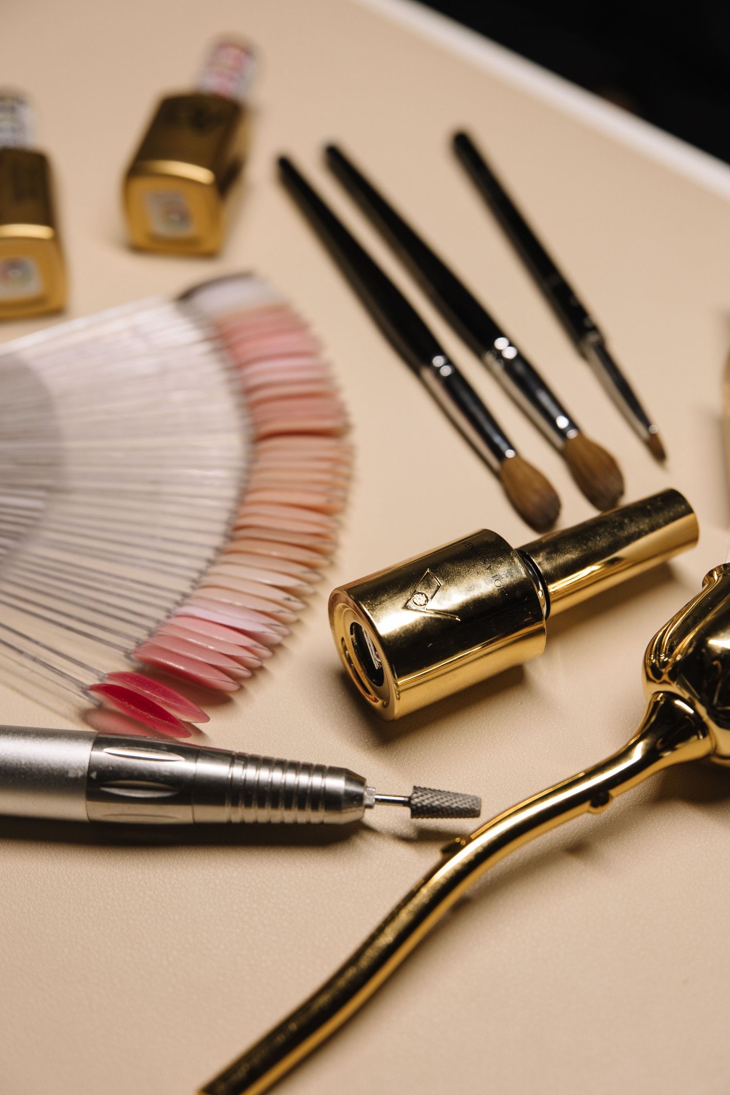 Professional nail care tools including brushes, gold eyelash curler, and color swatches arranged on beige background.