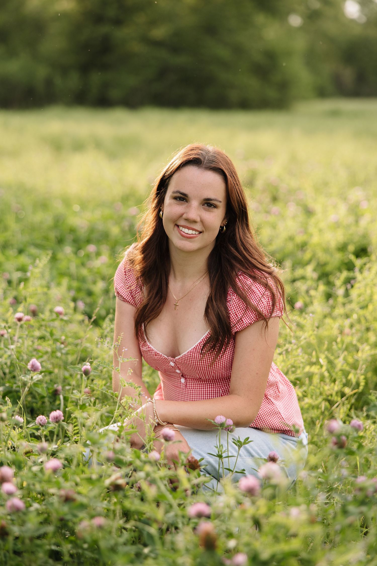 Smiling young woman with long brown hair sitting in a lush green field of wildflowers wearing a pink checkered top.