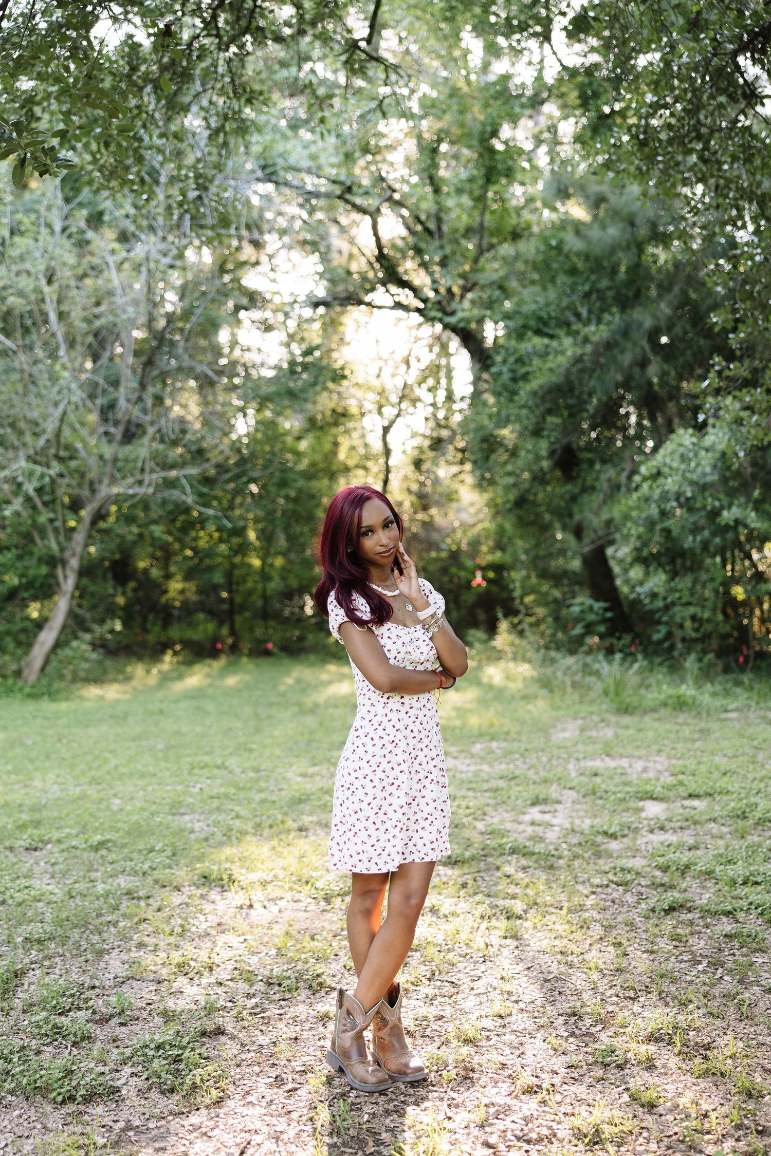 Young woman in white floral dress with red hair posing outdoors on a sunlit path surrounded by lush green trees.