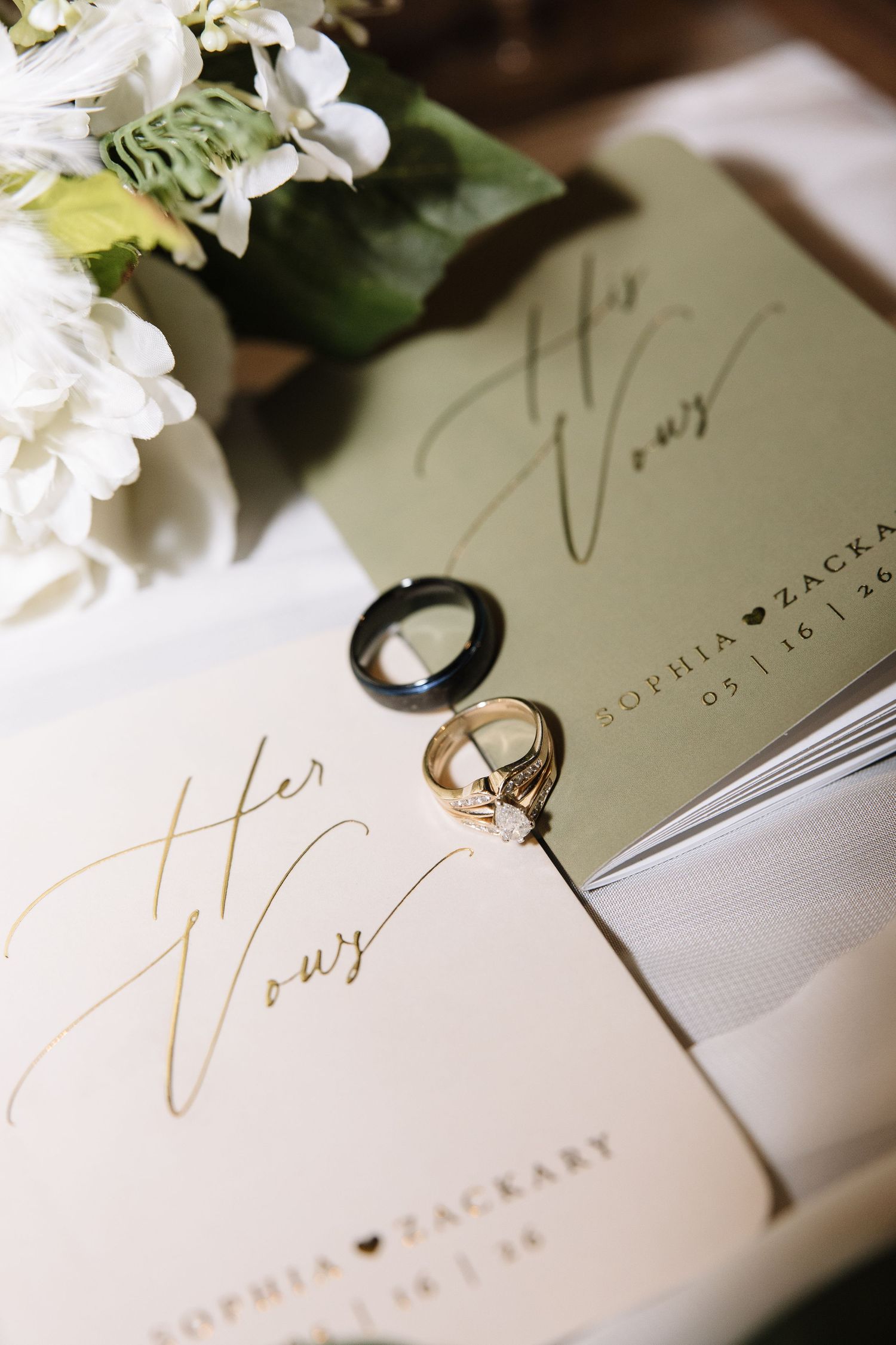 Wedding vow booklets for Sophia and Zackary with gold and black rings on elegant cream and sage paper.