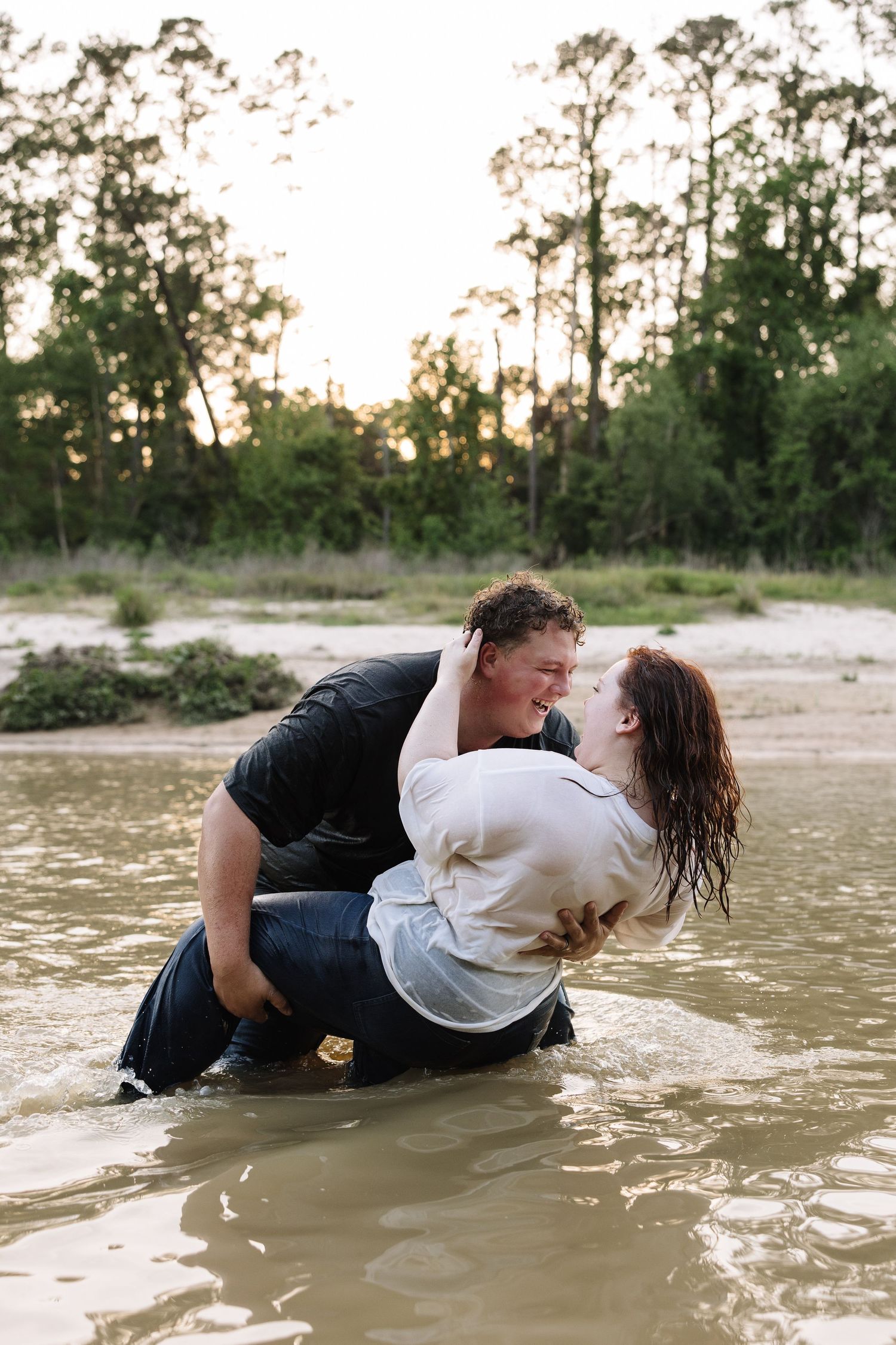 Couple sharing a romantic moment while wading in shallow water, surrounded by lush trees at sunset.