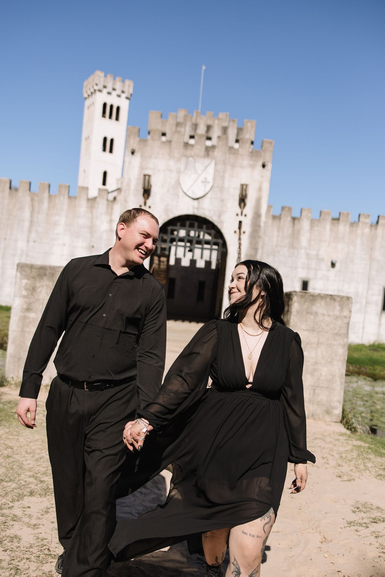 Couple in matching black outfits holding hands and smiling in front of a white castle on a sunny day.