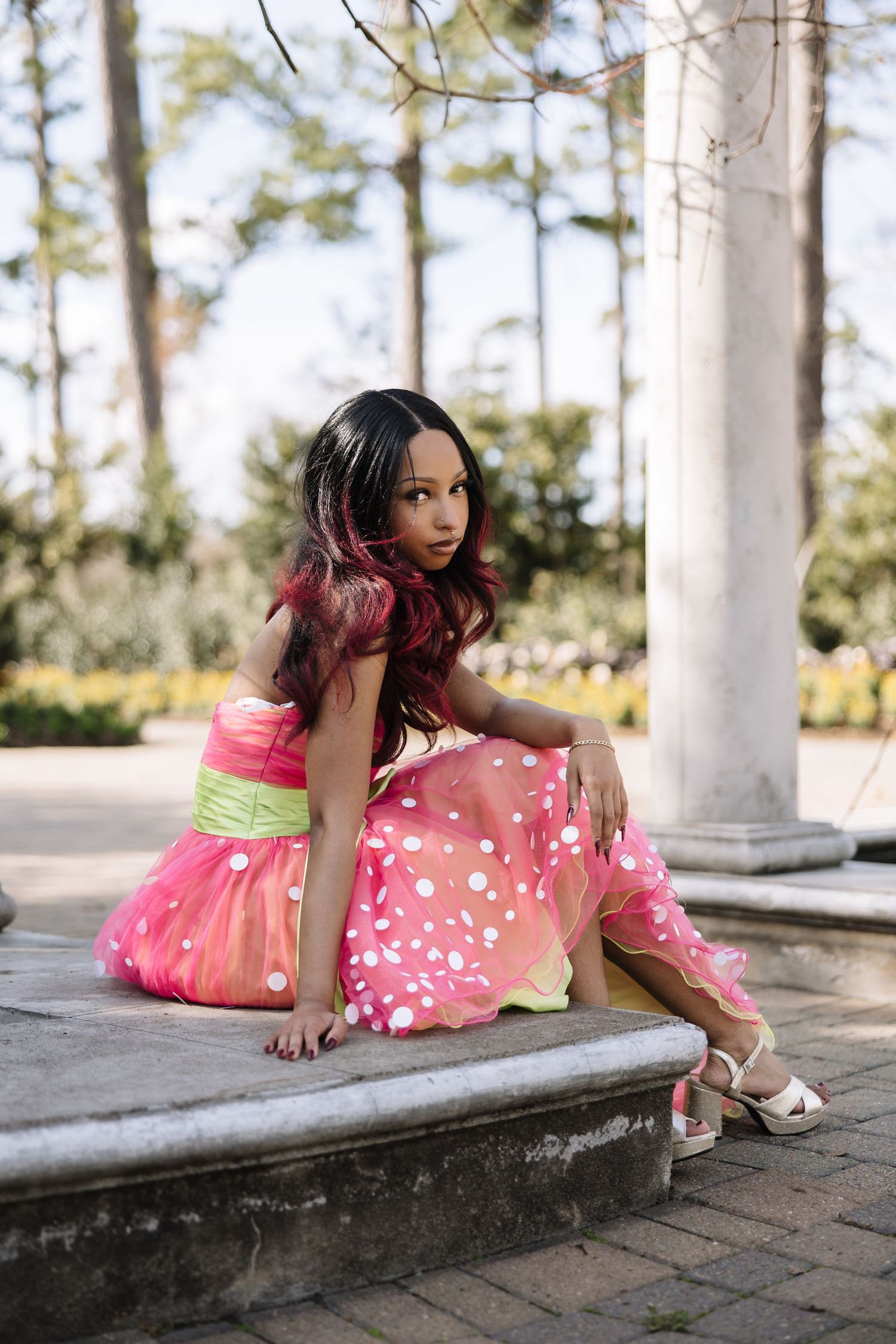 Young girl in pink polka dot dress with green sash sits on stone steps in a garden setting.